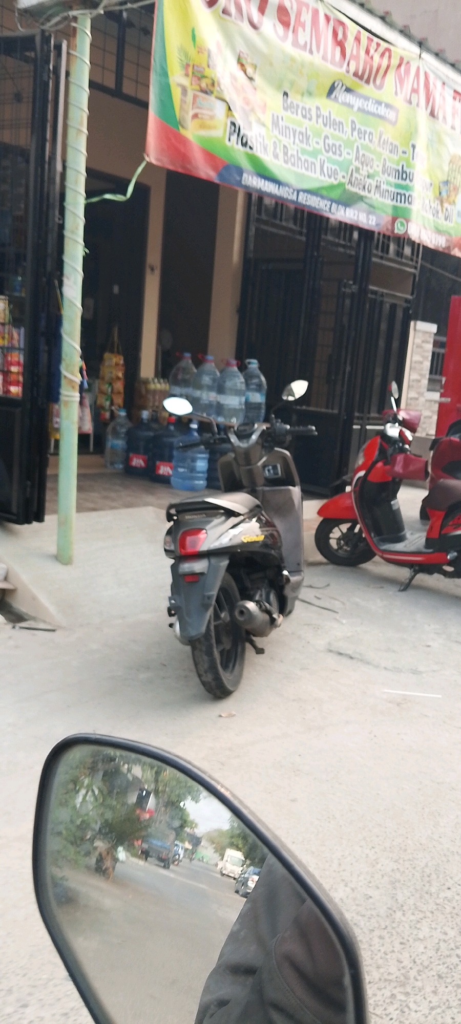 The image captures a street scene in Satriamekar, Indonesia. In the foreground, a motorcycle's rearview mirror shows a blurred reflection of a road with several vehicles, including a white van and a dark car, along with trees and buildings. The main part of the image focuses on two parked scooters. A black scooter is positioned in the center, viewed from the rear, with its exhaust pipe visible. To its right, a red scooter is parked. Behind the scooters, several large blue water jugs are stacked, and a banner with Indonesian text, partially obscured, hangs above. The setting appears to be an outdoor area in front of a building.