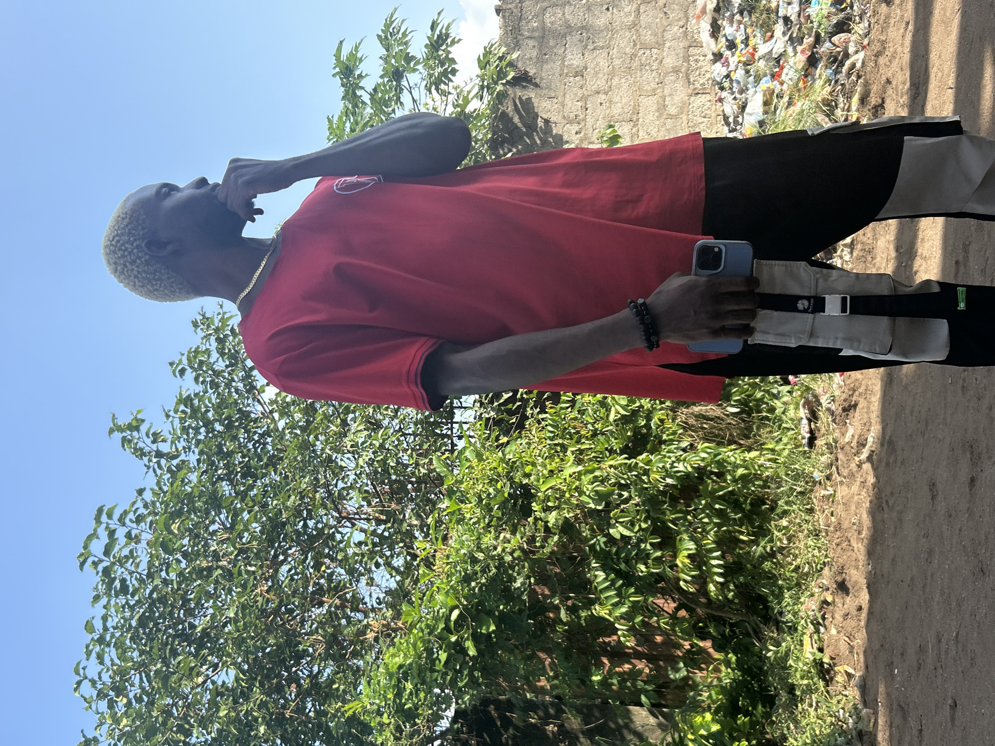 In the city of Ihiagwa, Nigeria, a Black male stands outdoors, facing right, with his left hand touching his chin. He has dyed blonde hair and wears a red t-shirt, black and beige cargo pants, and a beaded bracelet on his left wrist. He holds a blue smartphone in his right hand, which is tucked into a utility pouch on his pants. The background features lush green foliage to the left, a stone wall behind him, and a pile of trash to the right. The ground is dirt and there is evidence of scattered debris. The sky is clear and blue.