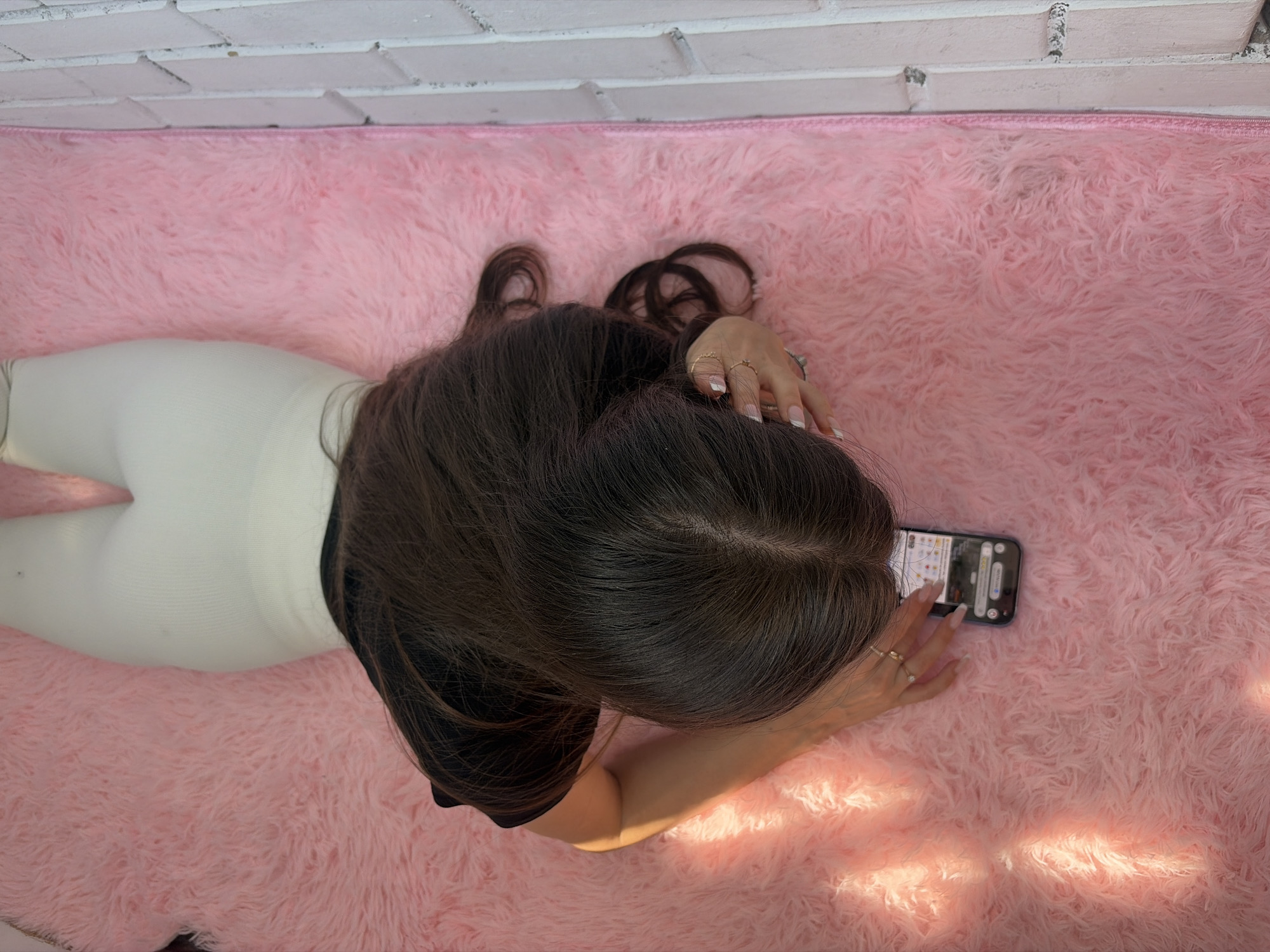A woman lies on her stomach on a pink, shaggy rug with her back facing the camera. She is wearing white leggings and a black top. She is holding a smartphone in her hands and appears to be looking at the screen. Her long, dark hair is spread out on the rug. The background is a white brick wall. The location cannot be determined.