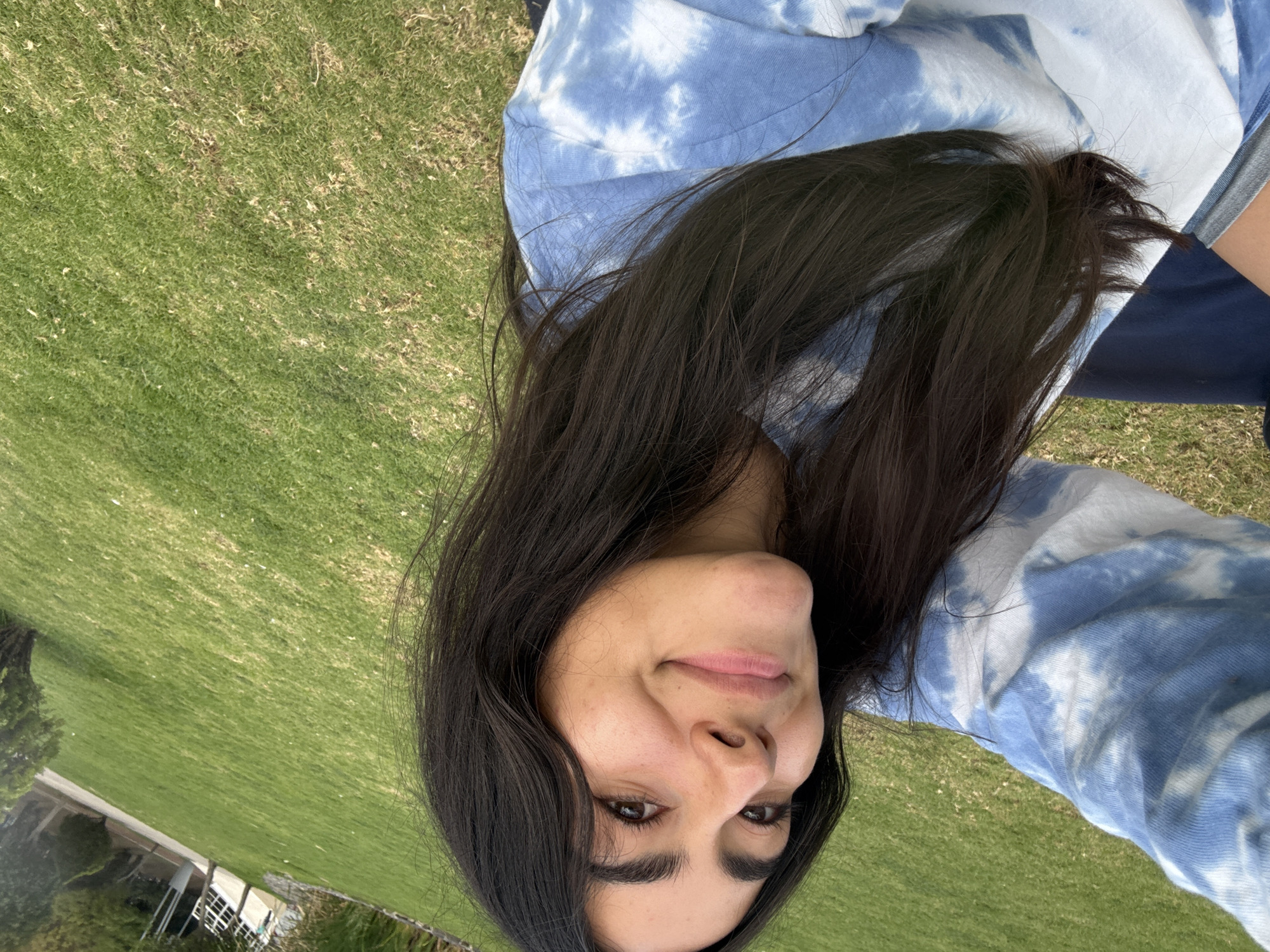 A young woman with long, dark hair is centered in the frame, smiling subtly. She is wearing a blue and white tie-dye hoodie. The background consists of a large, green lawn. A building is visible in the distant left corner. The location cannot be determined from the information provided in the image.