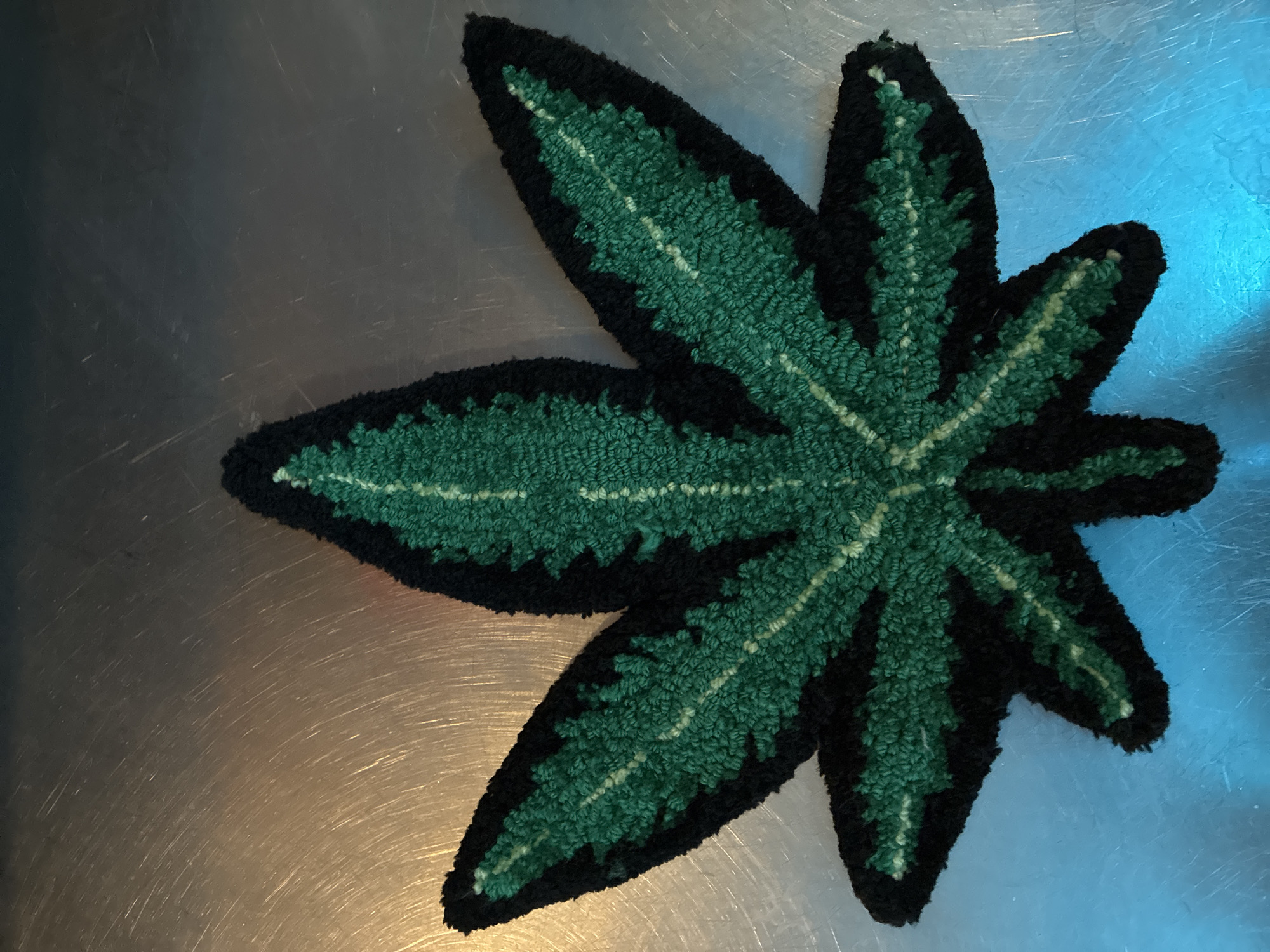 This image displays a handcrafted textile item, shaped distinctly like a cannabis leaf, positioned on a reflective metallic surface. The leaf features a textured, looped pile, characteristic of techniques like punch needle or rug hooking. Its design incorporates various shades of green; a dark green forms the main body of the leaf, while lighter green lines delineate the veins, all outlined by a thick border of black yarn.
The metallic surface beneath the leaf appears to be brushed or scratched, exhibiting different color reflections across its area. The upper left portion of the background reflects a darker, muted tone. Towards the lower left, the surface reflects a warm, golden hue, suggesting a nearby light source or environmental color. Conversely, the upper right section of the background displays a cool, vivid blue reflection.
The image is a still life, with no people or activities taking place. There is no visible text. The time of day and weather cannot be determined from the photograph. While the provided context indicates the location as Barcelona, Spain, there are no specific visual cues within the image itself to confirm this geographical detail.