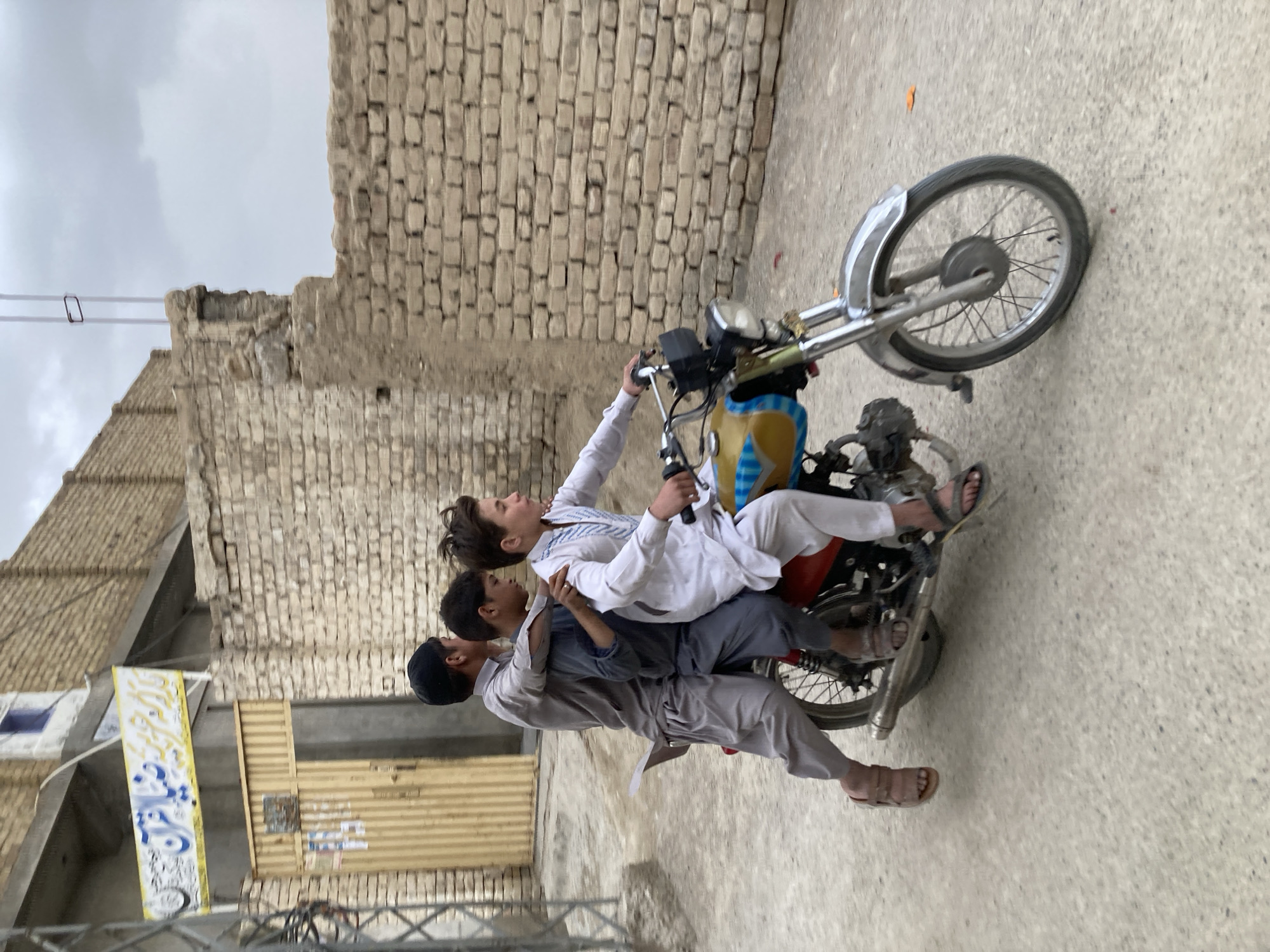 Three boys ride a motorcycle in Quetta City Tehsil, Pakistan. The boy driving is wearing a white shirt and pants. The other two boys are wearing gray clothing. They are on a paved road next to a brick building. There is a yellow and blue banner on a nearby building. The sky is overcast.