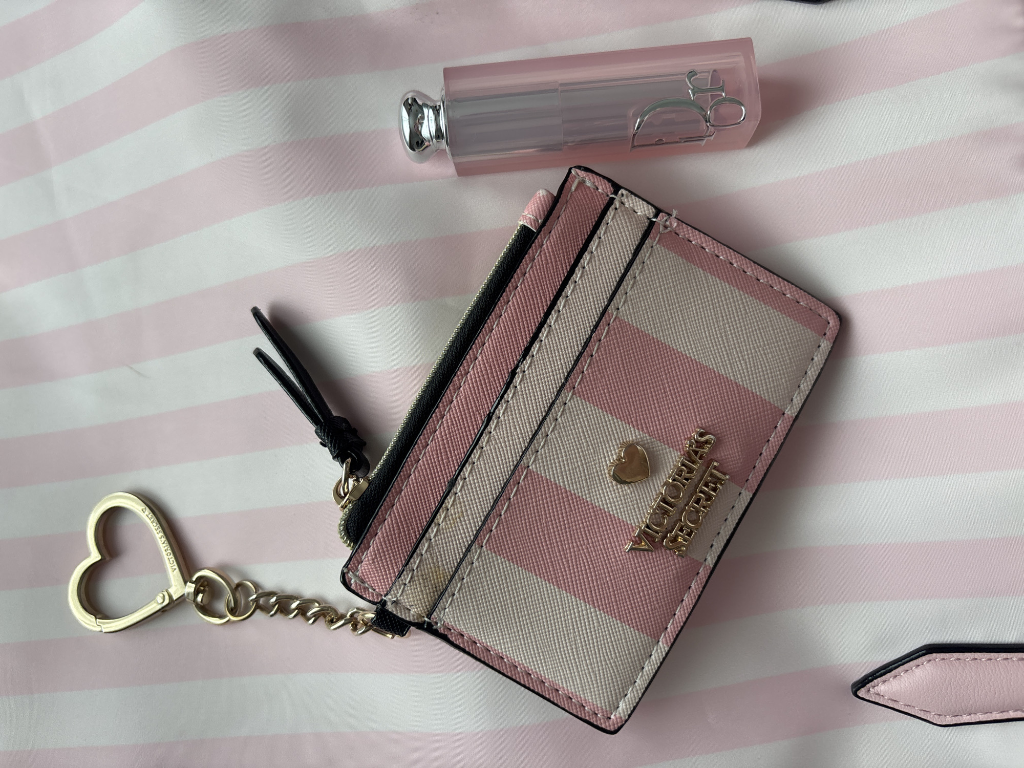 The image shows a pink and white striped Victoria's Secret cardholder with a gold heart keychain attached, a Dior lip maximizer, and a background of pink and white stripes. The cardholder is the most prominent object, featuring a striped pattern and the "Victoria's Secret" logo in gold lettering. The Dior lip maximizer is positioned above the cardholder. The entire scene is set against a background of pink and white stripes. There is no indication of the location in a specific city.