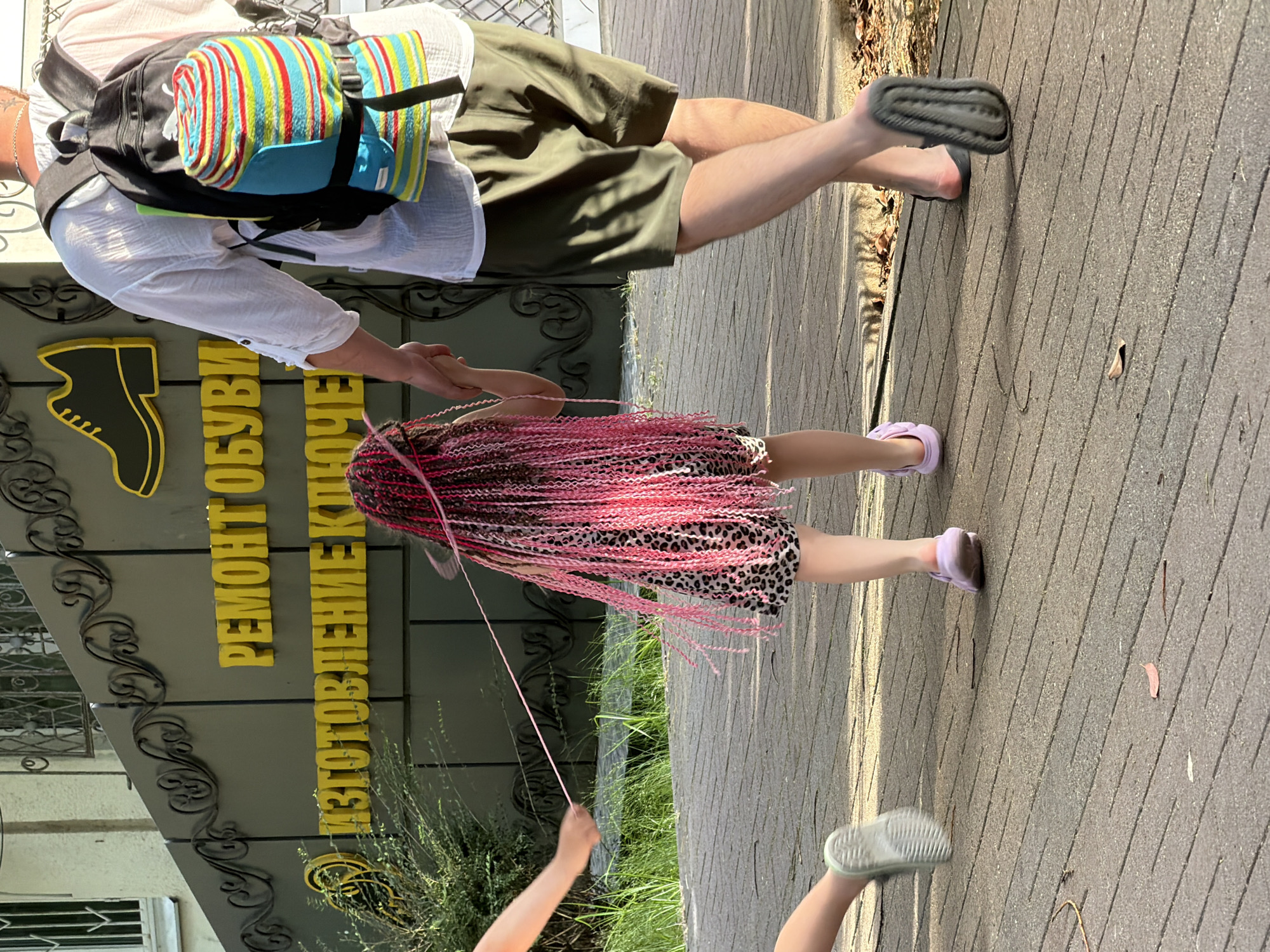 The image shows two people on a paved walkway in front of a building. The person in the foreground, from the knees down, is a child with long, pink braided hair wearing a leopard-print dress and pink sandals. They are holding a thin, light pink string. The other person is an adult, whose legs are visible from the knees down. They are wearing khaki shorts and dark grey slides, with a backpack with a colorful rolled-up blanket on their back. The adult is holding the child's hand.
The setting appears to be outdoors, likely in the daytime, given the shadows cast on the walkway and the bright sunlight. The building behind them has a sign with yellow lettering that reads "РЕМОНТ ОБУВИ ИЗГОТОВЛЕНИЕ КЛЮЧЕ". This translates to "SHOE REPAIR, KEY MAKING". The architecture of the building's entrance, with decorative metalwork and a shoe logo, suggests it is a shop. There is some greenery and trees visible, indicating a possibly pleasant weather day. The overall mood seems casual and everyday.
