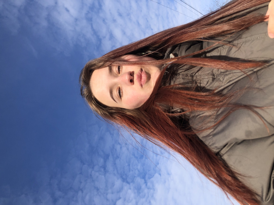 A close-up, upward-angled outdoor photograph features a young female individual against a clear blue sky interspersed with scattered white clouds, taken in the city of Фоки, Russia. The subject has long, straight, reddish-brown hair, parted centrally and falling over her shoulders and chest. Her face is angled slightly upwards, with her eyes partially squinted and her mouth puckered. She is wearing a dark grey or olive-green puffer-style jacket. A single, long, thin, chain-like earring is visible on her left ear. Strong, direct sunlight illuminates the subject's face from the right. A faint, dark linear element, possibly a utility line or tree branch, is discernible in the lower right background.
