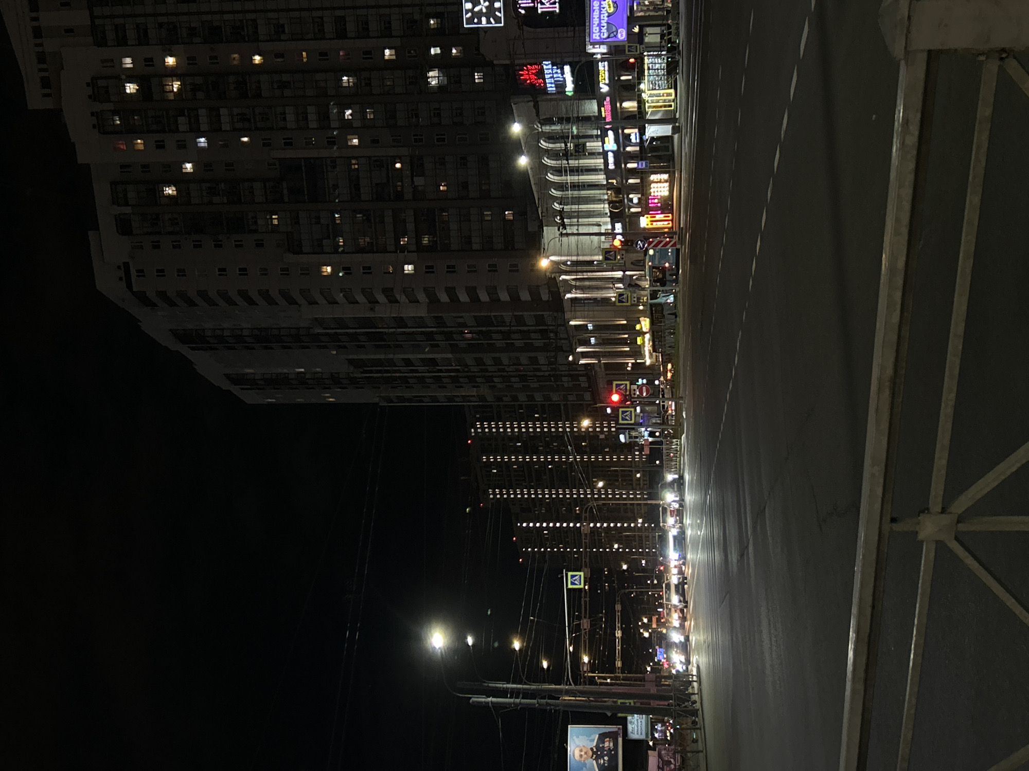 The image displays a vibrant urban street scene at night, appearing rotated 90 degrees clockwise. When viewed correctly, a tall, modern building dominates the left side, its numerous windows softly glowing from interior lights.
The multi-lane asphalt road with dashed white lines stretches into the distance on the right, illuminated by an array of streetlights and the headlights and taillights of vehicles, suggesting ongoing traffic. Along the right side of the street, commercial buildings are adorned with a dense collection of neon and backlit signs. Visible Cyrillic text includes "ДОЧКИНЫ СКИДКИ" (Dochkiny Skidki – "Daughter's Discounts") on a purple sign and "ПИВНУШКО" (Pivnushko – "Pub") on a prominent red sign. A clock-like sign displaying "1K" or "IK" is visible near the top of the tall building.
Utility poles with intricate wiring crisscross the frame. A red traffic light is clearly visible, directing street flow. In the lower right corner, a billboard features the face of a man, possibly in a uniform, with partially discernible Cyrillic text such as "Лен С.". A metal barrier or railing partially frames the right edge of the image. The dark night sky highlights the artificial illumination, creating a dynamic cityscape typical of Saint Petersburg, Russia.