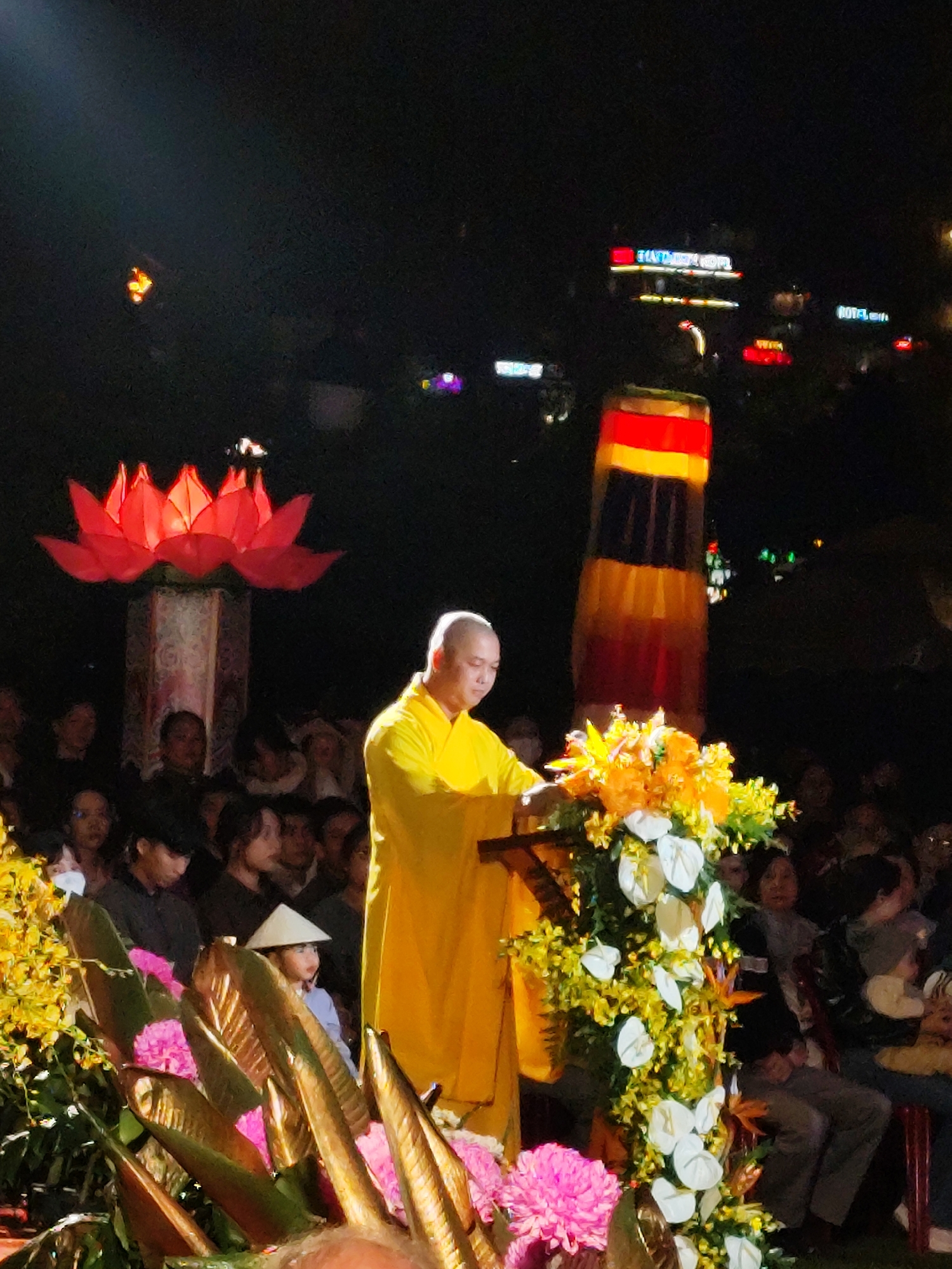 At night in Da Lat, Vietnam, a bald monk in a yellow robe stands behind a lectern adorned with yellow and white flowers, giving a speech to a large, diverse audience. Behind the monk is a tall, cylindrical decoration with horizontal bands of red, yellow, brown, and black. To the left, a large illuminated red lotus flower stands atop a decorated pillar. A child in a conical hat sits in the front row. In the distance, brightly lit buildings display the words "DA LAT HOTEL" and "ROYAL." Other lights and buildings are visible in the background. The scene is crowded, with many faces visible in the audience.