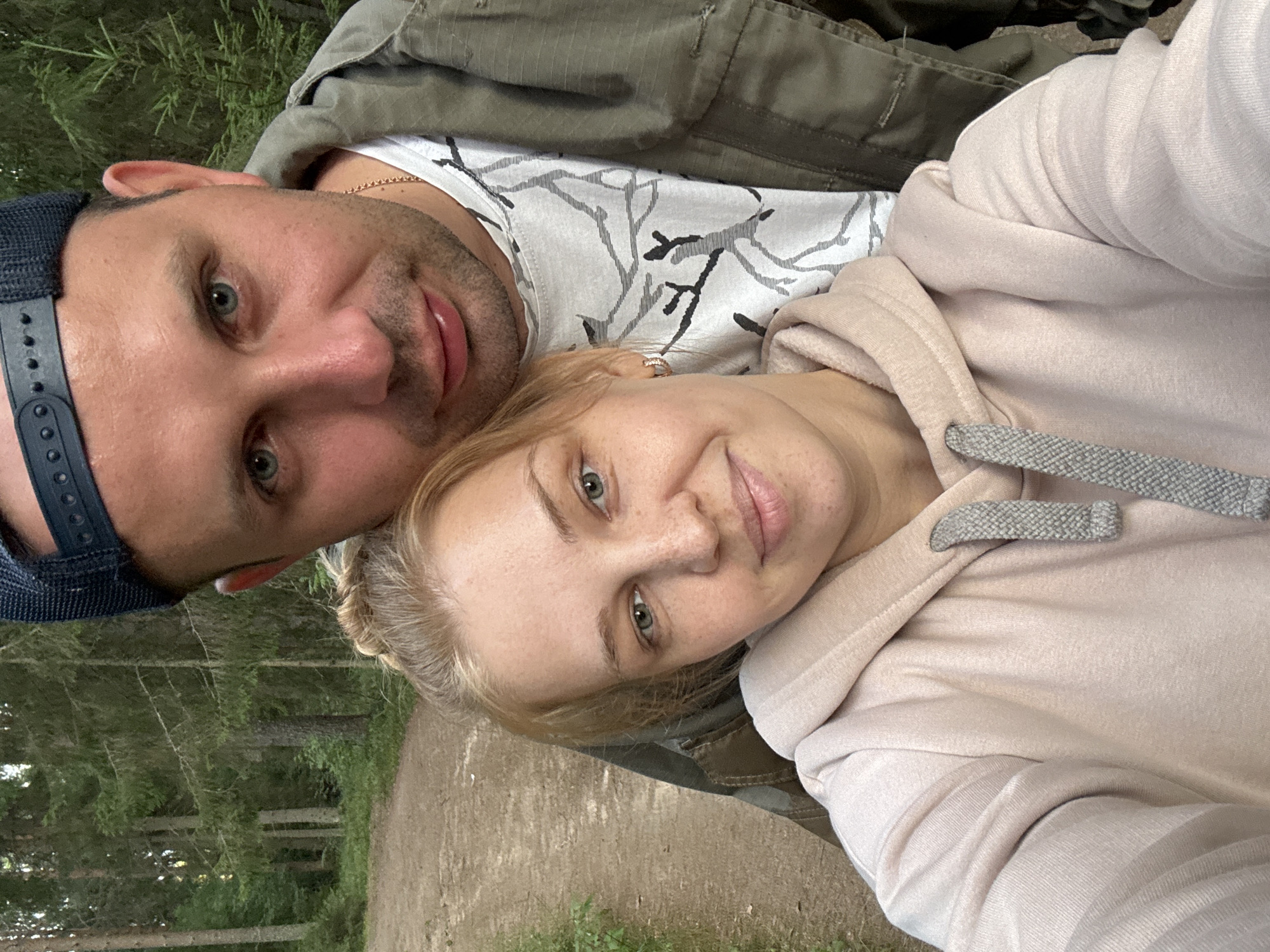 In the city of Anteplevo (Okhotinskoe S/P), Russia, a close-up, selfie-style photograph captures a Caucasian couple standing outdoors on a dirt path surrounded by a forest. The woman, in the foreground, has blonde hair pulled back in a bun and is wearing a light pink hoodie with grey drawstrings. She has light skin with freckles, blue-green eyes, and is smiling. The man is positioned behind and slightly to the right of the woman, leaning his head towards her. He has short brown hair, blue eyes, and is wearing a black baseball cap, a white t-shirt with a grey abstract pattern, and an olive green jacket. The background consists of tall evergreen trees and the path, suggesting a natural, wooded environment. The lighting indicates it is daytime.