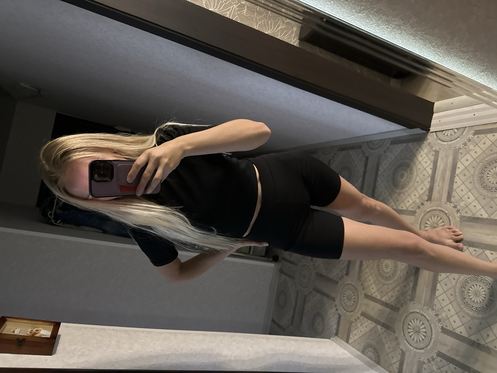 The image shows a blonde woman in black athletic wear, taking a mirror selfie. She is standing in what appears to be an indoor hallway, possibly in an apartment. The flooring is tiled with a repeating geometric pattern. To the left, a small wooden cabinet with a framed picture is mounted on the wall. To the right, a doorway with a dark frame leads to another room. The overall lighting is dim, with some light coming from an unseen source. The scene does not contain any specific indicators to place it within the city of Energetik, Russia.