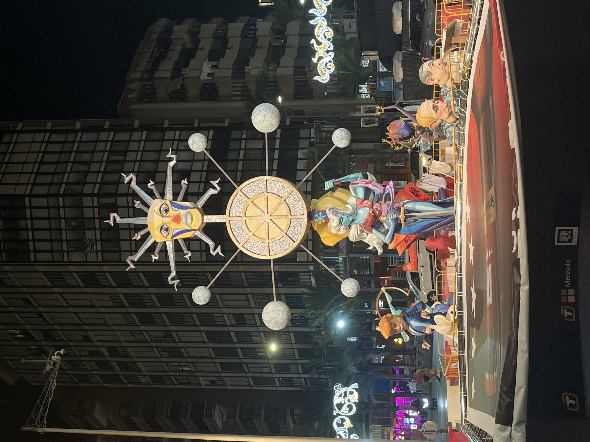 The image captures a nighttime scene in Alicante, Spain, featuring a large, elaborate artistic display. Dominating the foreground is a satirical monument constructed of brightly colored materials, likely part of a festival such as Las Fallas. The monument depicts several caricatured figures, including one resembling Donald Trump holding a bow and arrow, and another that could be Vladimir Putin. A stylized, humanoid figure with blue skin and flowing hair stands prominently, holding a sword. Above these figures, a large, circular structure with radiating spokes resembles a sun or a celestial body, with a face at its center and spherical elements extending outwards. In the background, tall, modern buildings with many windows are visible, along with palm trees and streetlights, indicating an urban setting. Decorative lights are strung across the street, and several vehicles, including a white van and cars, are present. The overall atmosphere is festive and vibrant, characteristic of a public celebration at night.