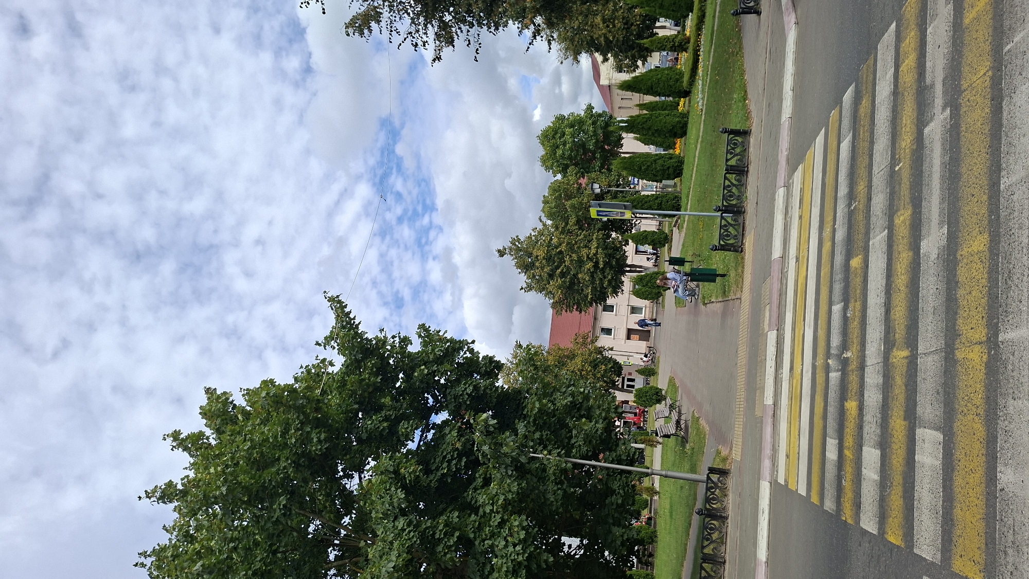 This image captures a street scene in Pastavy, Belarus, featuring a prominent pedestrian crossing in the foreground. The crossing is marked with alternating yellow and white stripes. Beyond the crossing, a pathway leads into a park-like area with green grass and trees. Several park benches are visible, and a few individuals are seated or walking in the park. Buildings with light-colored facades and red roofs line the background. The sky above is partly cloudy. The scene appears calm and ordinary, depicting a typical day in a public space.