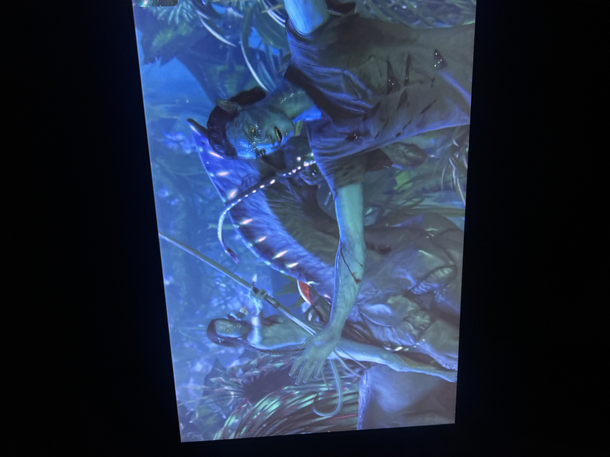 The image displays a vibrant, low-light scene from the film *Avatar*, captured from a screen. The primary subject is a blue-skinned Na'vi character, likely Jake Sully in his avatar form, depicted in a dynamic, diagonal orientation within the frame. His head is positioned towards the upper right, and his body extends downwards towards the lower left.
The Na'vi has characteristic glowing yellow eyes, pointed ears, and intricate facial patterns. He is wearing a dark, short-sleeved tunic or shirt, which shows some red marks, possibly blood or battle wounds. A glowing, beaded adornment is visible around his neck and chest. His mouth is slightly open, and his expression suggests intensity, effort, or distress. His left arm is outstretched towards the bottom of the frame, revealing more red markings.
The background depicts a dense, alien jungle, typical of Pandora, bathed in bioluminescent blues, greens, and purples. This suggests a nighttime or deep-forest setting, with the glowing flora creating a magical yet potentially dangerous atmosphere. Partially visible below the main Na'vi, and somewhat obscured, is another figure or element, with a hand visible and a long, slender object, possibly a spear or staff, nearby. The scene conveys a moment of action, struggle, or peril.
No visible text is discernible within the image content. The image itself appears to be a photograph of a screen, indicated by the surrounding black borders and the slight sheen. This image was captured in Kazan, Russia.