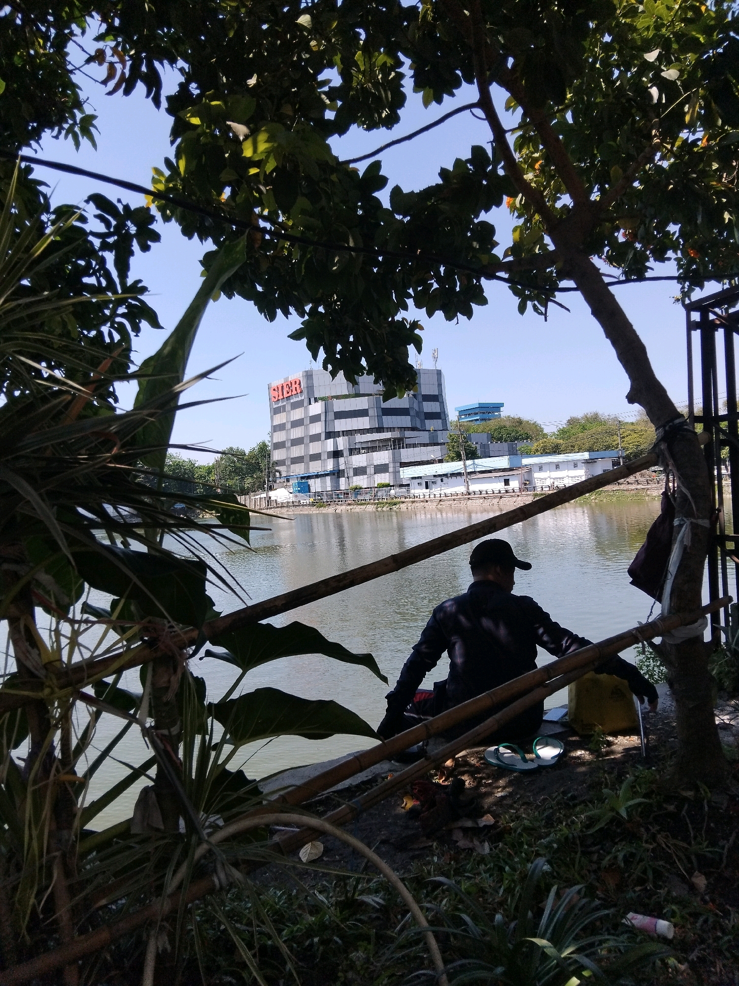 The image depicts a waterside scene in Surabaya, Indonesia, with a prominent building bearing the "SIER" logo in the background. In the foreground, a person wearing a black cap and dark clothing is seated on the bank of a body of water, facing away from the camera. Their arms are resting on a bamboo railing, and they appear to be looking out at the water. Next to them, a yellow bag and a pair of flip-flops are visible on the ground. The area is surrounded by lush greenery, with trees and foliage obscuring parts of the view. The sky is clear and blue. The building in the background is multi-story with a checkered facade and appears to be an industrial or office complex. The body of water is calm and reflects the surrounding environment.