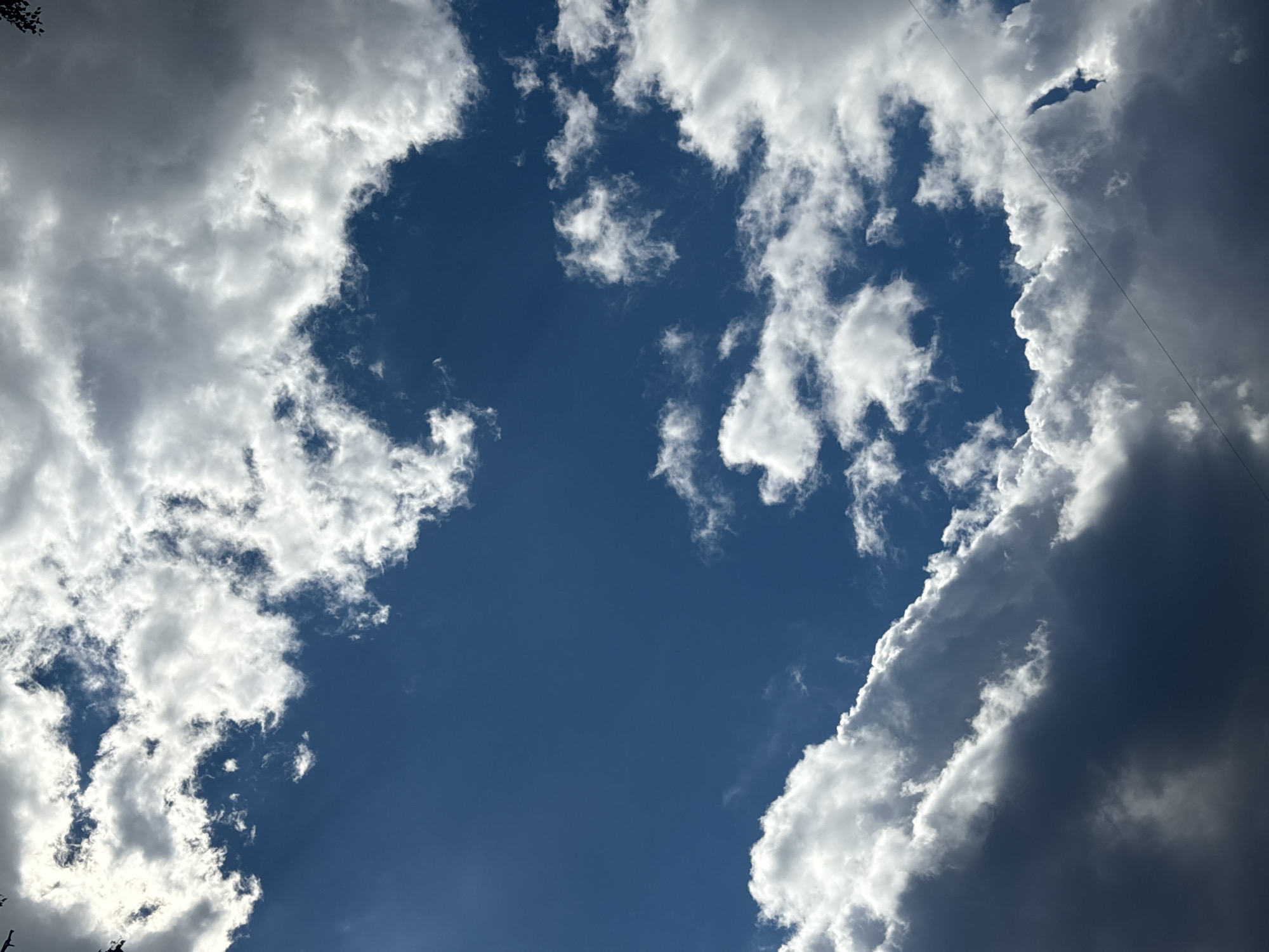 This media file captures a dynamic sky filled with a mix of bright white and dark gray clouds against a deep blue background. The clouds are textured and voluminous, suggesting a partly cloudy or potentially stormy day. Sunlight is filtering through and illuminating some of the clouds, creating a dramatic contrast between light and shadow. One edge of the frame shows a dense, dark cloud formation, while other areas feature wispy, bright clouds. There are faint, thin lines visible crisscrossing the sky, likely representing power lines or wires, suggesting an outdoor, possibly rural or suburban, setting. The overall impression is one of natural beauty and the changing moods of the sky. The time of day appears to be daytime, given the bright sunlight and clear blue sky. No people, specific objects, or visible text are present in the image.