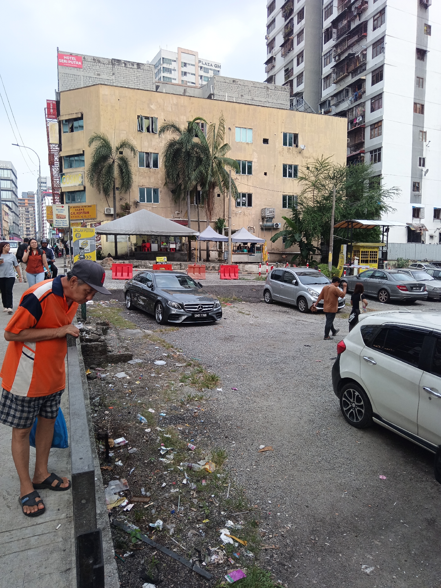 The image depicts an outdoor scene in Kuala Lumpur, Malaysia. A man wearing an orange polo shirt and checkered shorts is standing on a concrete sidewalk, leaning over a low wall and looking down at a debris-strewn area. A blue plastic bag is visible next to his feet. In the background, several cars are parked on a gravel lot. One car, a dark grey Mercedes-Benz, is prominently featured. Behind the parking lot are several buildings, including a tan-colored building with palm trees in front of it, and taller, modern apartment buildings. Various signs are visible on the buildings, including "HOTEL SERI PUTRA" and "PLAZA GM." The overall environment appears to be a somewhat gritty urban setting with accumulated litter on the ground.