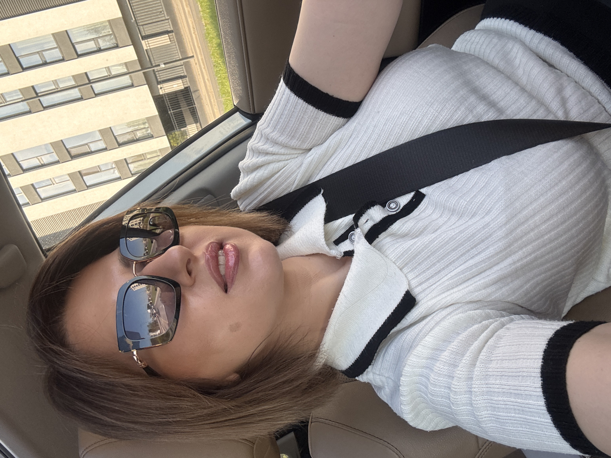 A woman is sitting in a car, wearing a white sweater with black trim and a seatbelt. She is wearing sunglasses. Outside the car window is a building with many windows, and greenery can be seen as well. Based on the architectural style of the building, this could be a location in Sofia, Bulgaria.