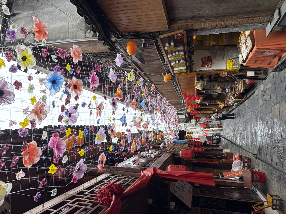 A narrow, stone-paved street in Furong, China, is depicted, appearing wet and sloping gently downwards into the distance under an overcast sky. The street is densely covered overhead by a white net adorned with numerous colorful, translucent artificial flowers in shades of pink, orange, yellow, blue, and purple. Traditional multi-story wooden buildings with dark timber facades flank both sides of the street.
On the left, buildings feature ornate wooden railings and large red fabric decorations, including a prominent red bow and several cylindrical red lanterns suspended at various heights. A small wooden chair and a red table with items in plastic bags are positioned near the building's entrance.
On the right, buildings house shops with open fronts displaying merchandise such as woven hats, baskets, and packaged goods on wooden shelves and tables. An orange tablecloth with "Free Taste" in English and Chinese characters is visible. Multiple red lanterns and orange pumpkin-shaped decorative lights are affixed to the building facades and shopfronts.
In the mid-ground, a lone individual wearing dark trousers and a light-colored top with a backpack is walking away from the viewer, moving further down the street. Additional red lanterns are visible lining the street further back. The overall scene suggests a commercial or pedestrian area with festive decorations.