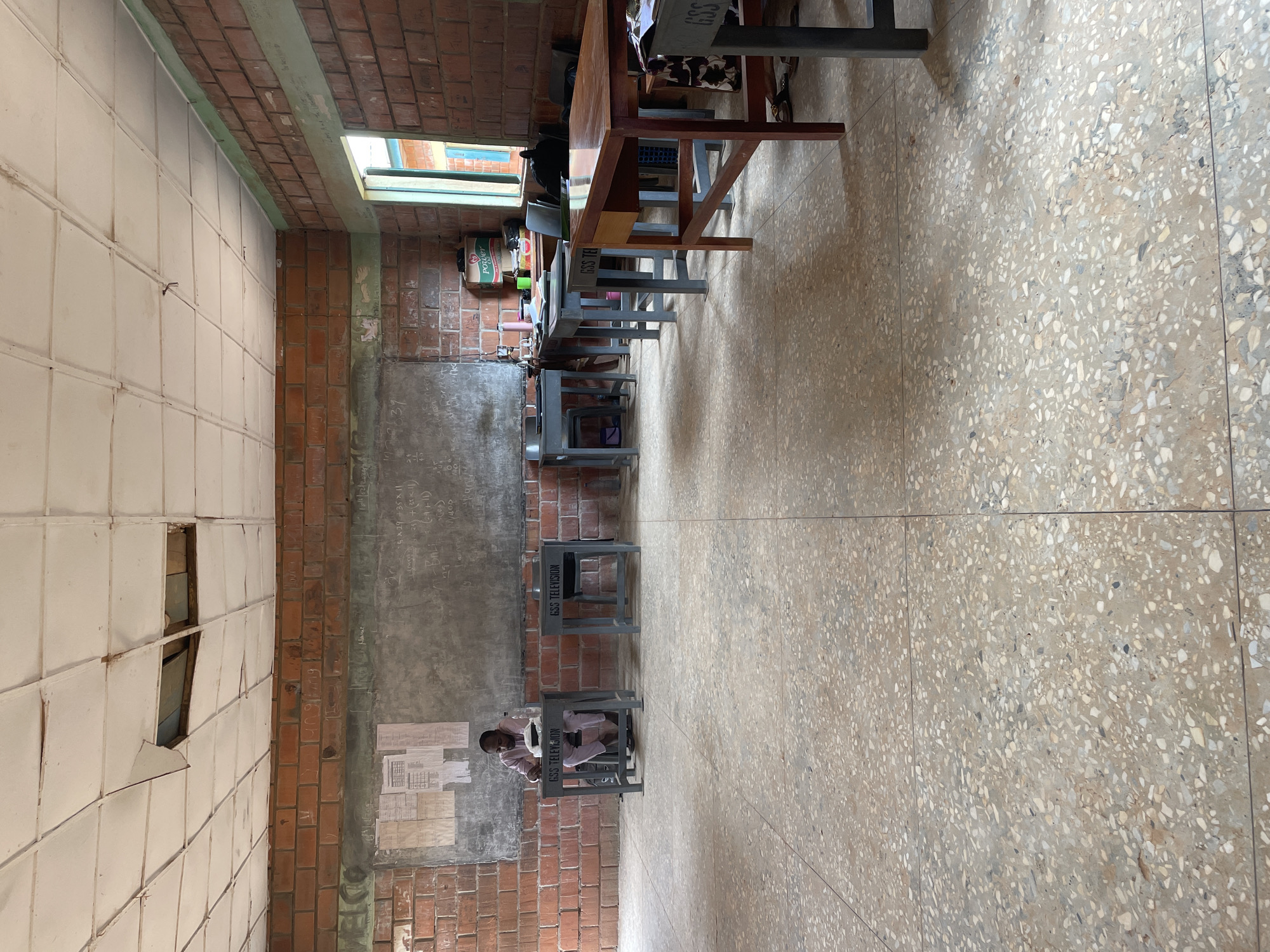The image depicts an interior classroom scene, likely in the city of Television, Nigeria. The room features rows of desks and chairs, some of which have "GSS TELEVISION" inscribed on them. A young person is sitting at one of the desks, facing the viewer. The walls are made of brick, with a section of tiled wall showing damage. A chalkboard is visible with writing on it. The floor is a tiled surface with a speckled pattern. Overhead, there's a narrow window providing natural light.