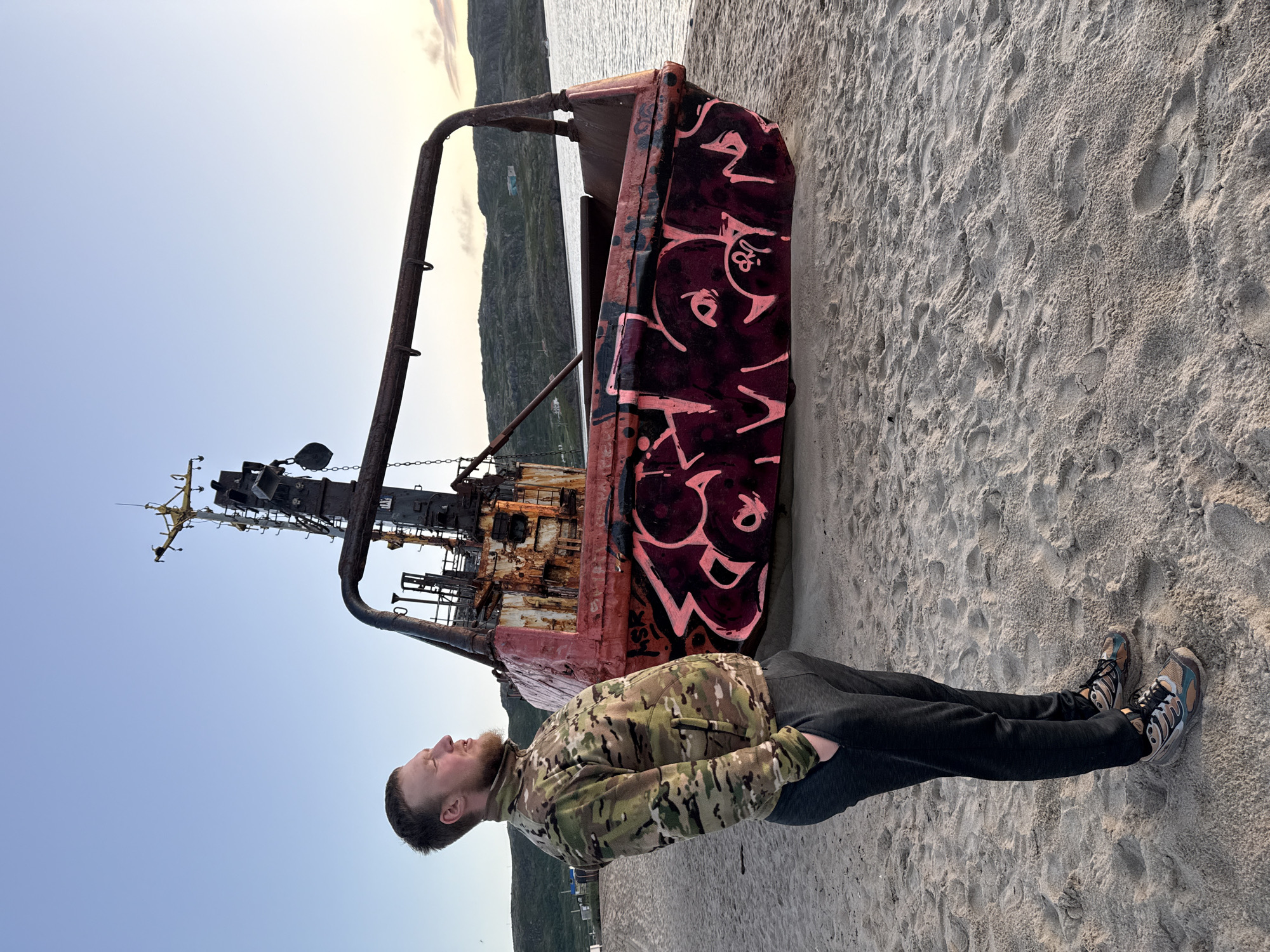 A man stands on a sandy beach in Teriberka, Russia, looking to his left. He is wearing a camouflage jacket, dark pants, and athletic shoes. Behind him is a rusty, dilapidated vessel covered in pink graffiti. The vessel appears to be beached on the shore. The sea is visible behind the vessel, and rolling hills with some sparse vegetation and buildings can be seen in the distance. The sky is clear and light blue, suggesting it is either early morning or late evening. The overall scene depicts a somewhat desolate and industrial seascape.