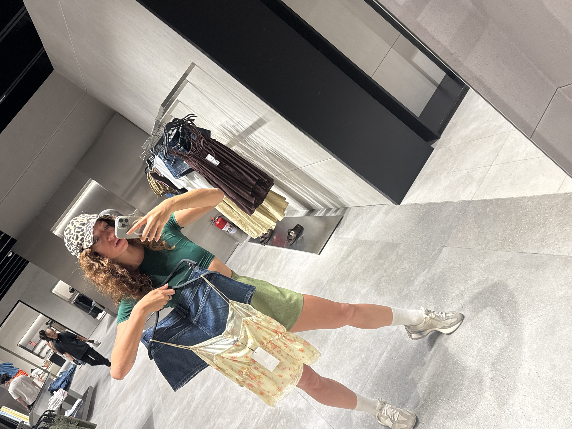A woman is taking a selfie in a clothing store. She is wearing a green top, green shorts, a leopard print hat, and white socks with sneakers. She is holding up two tops on hangers, one denim and one floral. The clothing store has clothing racks with items for sale. Other shoppers are visible in the background. The location is a clothing store, likely a Zara, in Hanoi, Vietnam.