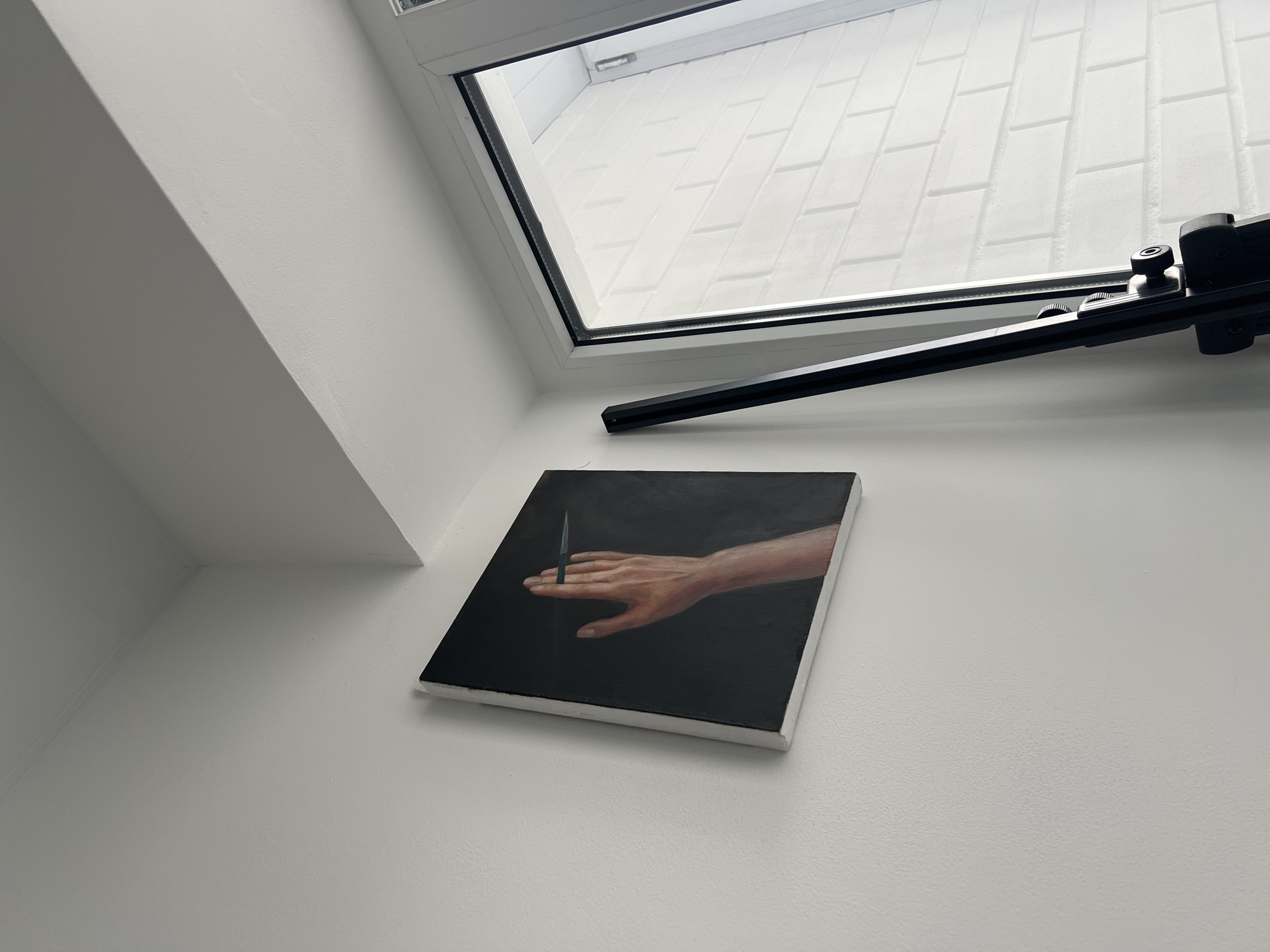 An oil painting of a human hand holding a pencil is displayed on a white wall. The painting has a dark background. To the right of the painting, a window with white brick is visible. A black easel is positioned in front of the window. The scene appears to be indoors in a room with white walls and a white ceiling.