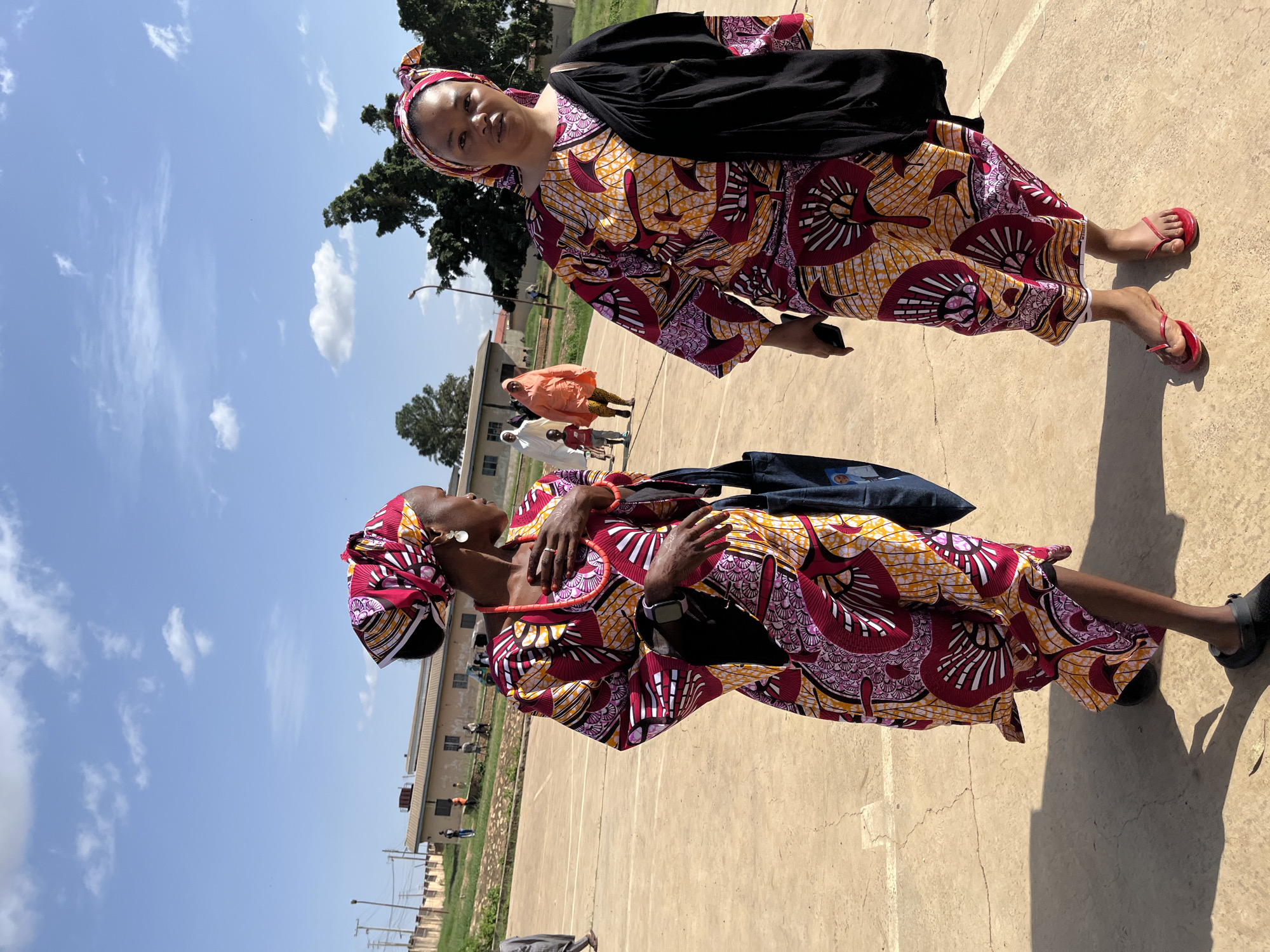 Two women in vibrant, patterned African attire are the primary subjects in the foreground of this outdoor image, likely taken in Jos, Nigeria. The woman on the left, with her headscarf matching her dress, turns to her right, her hand resting on her chest, adorned with jewelry. She carries a dark bag. The woman on the right faces forward, also wearing a matching headscarf and dress, with a dark shawl draped over her shoulder. She holds a dark object, possibly a phone, in her left hand.
The setting appears to be an open, paved area, possibly a courtyard or playground, with faint lines demarcating sections of the pavement. In the background, several other individuals are visible, including a child in a red shirt and at least two women in orange and white headscarves and dresses. A low building with a pitched roof and a wall are also present in the mid-ground, and further back, trees and utility poles suggest an urban or semi-urban environment. The sky is a bright blue with scattered white clouds, indicating a sunny day.