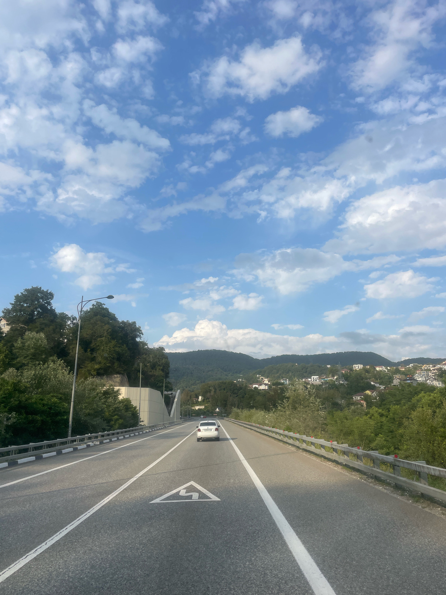 The image depicts a view from a car driving on a multi-lane highway. The road is asphalt with clear white lane markings, including a triangular sign with a stylized directional arrow. A white sedan is visible driving ahead in the right lane. On the left side of the highway, there is a metal guardrail and a line of trees, with a street light visible. Further left, a tunnel entrance with a curved, light-colored structure is present, and behind it, dense green trees and some houses are visible on a slope. The right side of the road also has a guardrail, and beyond it, the terrain slopes upwards, covered with lush green trees and dotted with numerous houses and buildings, suggesting a semi-urban or suburban residential area nestled on hillsides. The sky above is a bright blue with scattered, fluffy white clouds. The overall impression is a scenic drive through a hilly landscape with signs of habitation. The location is identified as the city of Razdolnoye, Russia.