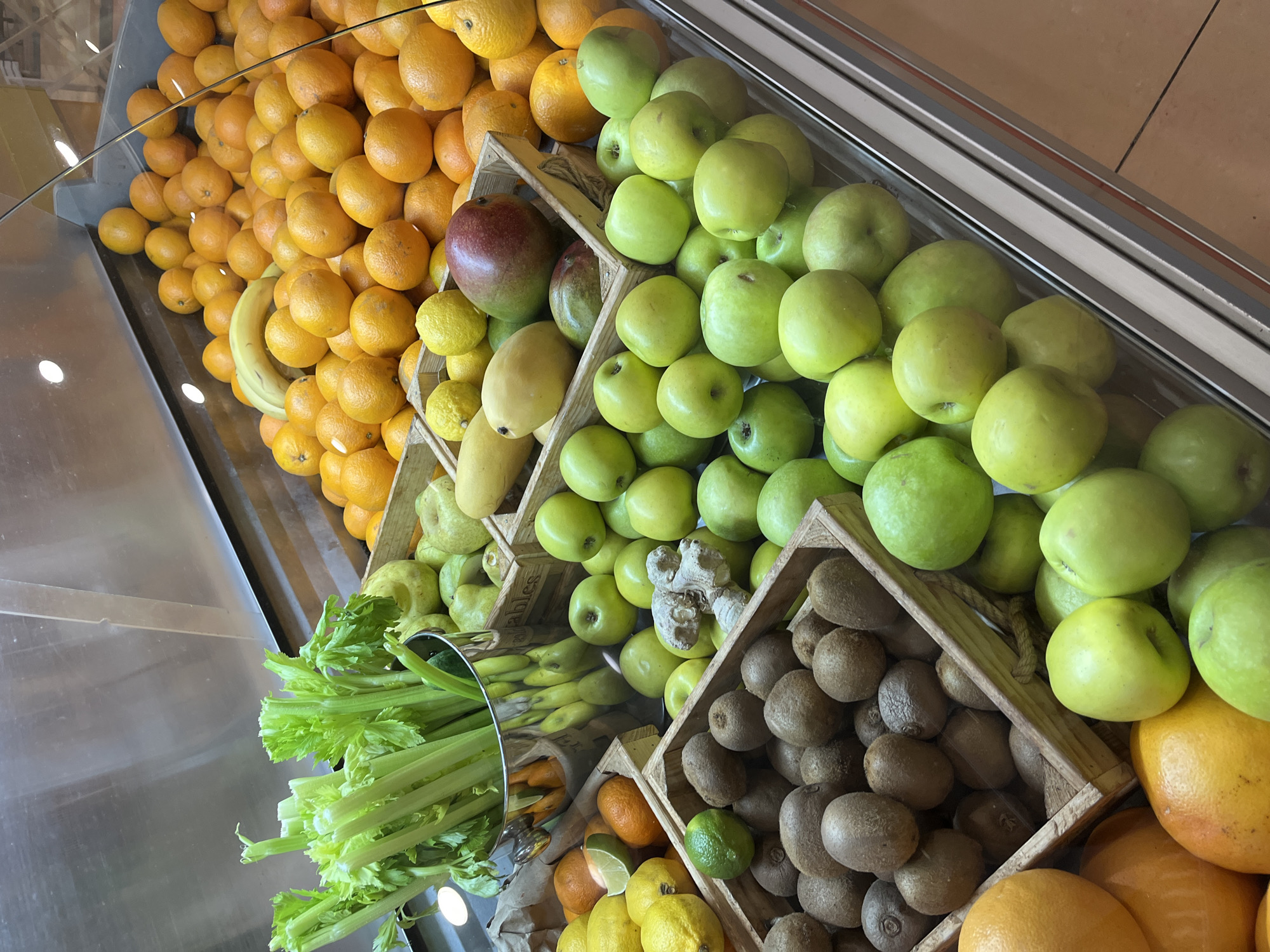 A high-angle shot depicts a display of produce in a refrigerated case, likely in a grocery store or market. The layout consists of multiple tiers. The top tier is almost entirely filled with a dense arrangement of oranges. A few yellow bananas are interspersed among the oranges. Below the oranges, there is a descending series of wooden crates. The crates contain various fruits and vegetables, including green apples, mangoes, lemons, limes, kiwis, pears, celery, and other citrus fruits. Some of the produce is spilling out of the crates. The back and side panels of the display are made of stainless steel. The location is unknown.