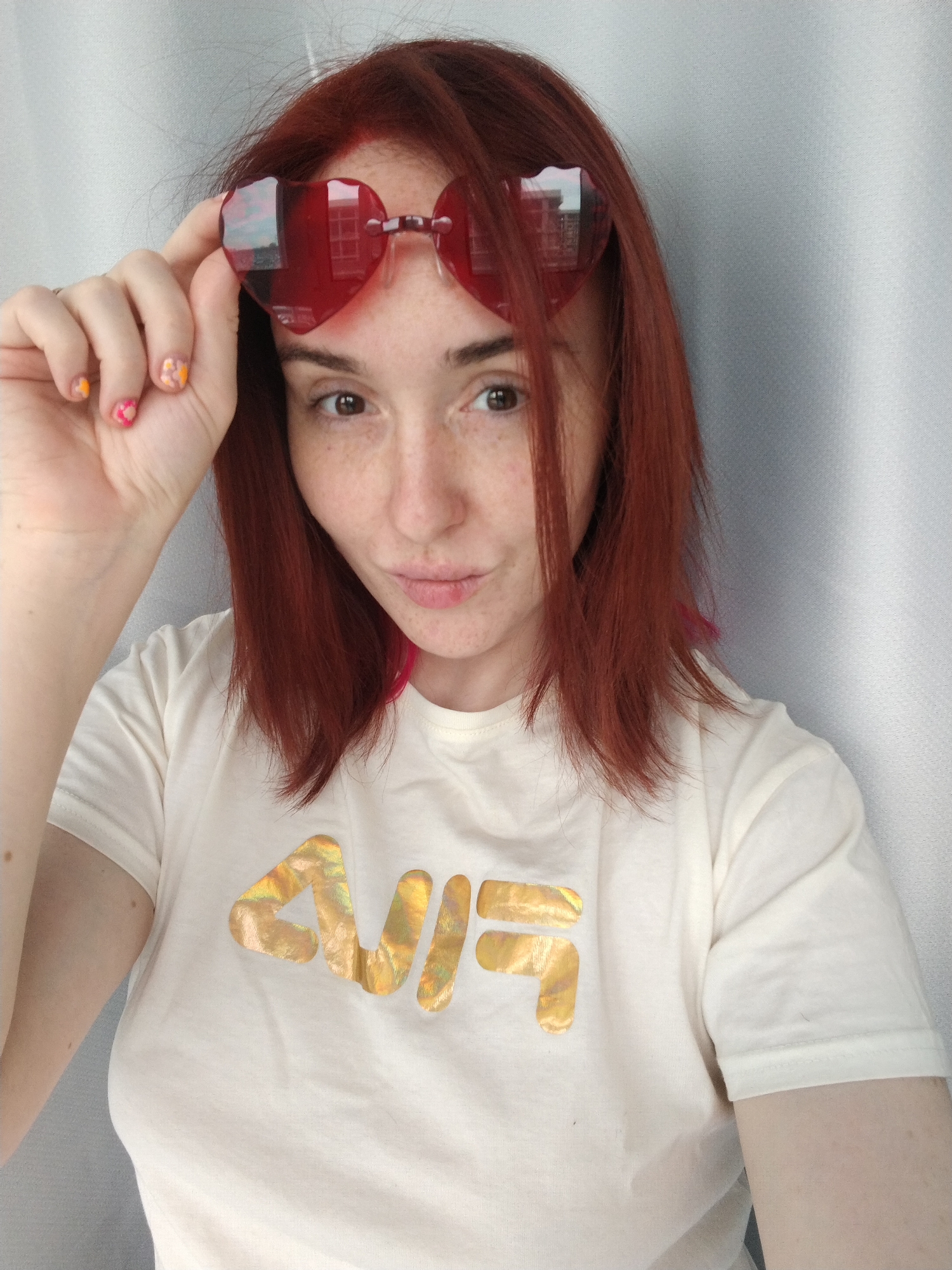 A female individual, appearing to be in her 20s or 30s, is depicted in a close-up, waist-up shot, facing the camera directly. She has reddish-brown, shoulder-length hair with subtle pink highlights visible on the right side (viewer's left). Her skin is fair, with visible freckles across her nose and cheeks. Her eyes are brown. She has a slight pout; a small lesion or blemish is present on her upper lip, slightly to the left of the philtrum.
She is wearing a cream-colored, short-sleeved t-shirt with a holographic gold "FILA" logo prominently displayed on the chest. Her right hand is raised, holding red, heart-shaped sunglasses by the bridge, positioned just below her eyes. The left lens of the sunglasses reflects a multi-story building with visible rectangular windows and a portion of the sky. Her fingernails are manicured, with the thumb and index finger displaying multi-colored, abstract floral or speckled designs in pink, orange, and yellow hues.
The background consists of a plain, light grey, textured fabric, consistent with a curtain or wall, indicating an indoor setting. No specific identifying landmarks are visible to determine a precise location within the city of Izhevsk, Russia, beyond the general indoor environment suggested by the background and the indistinct reflection in the sunglasses.