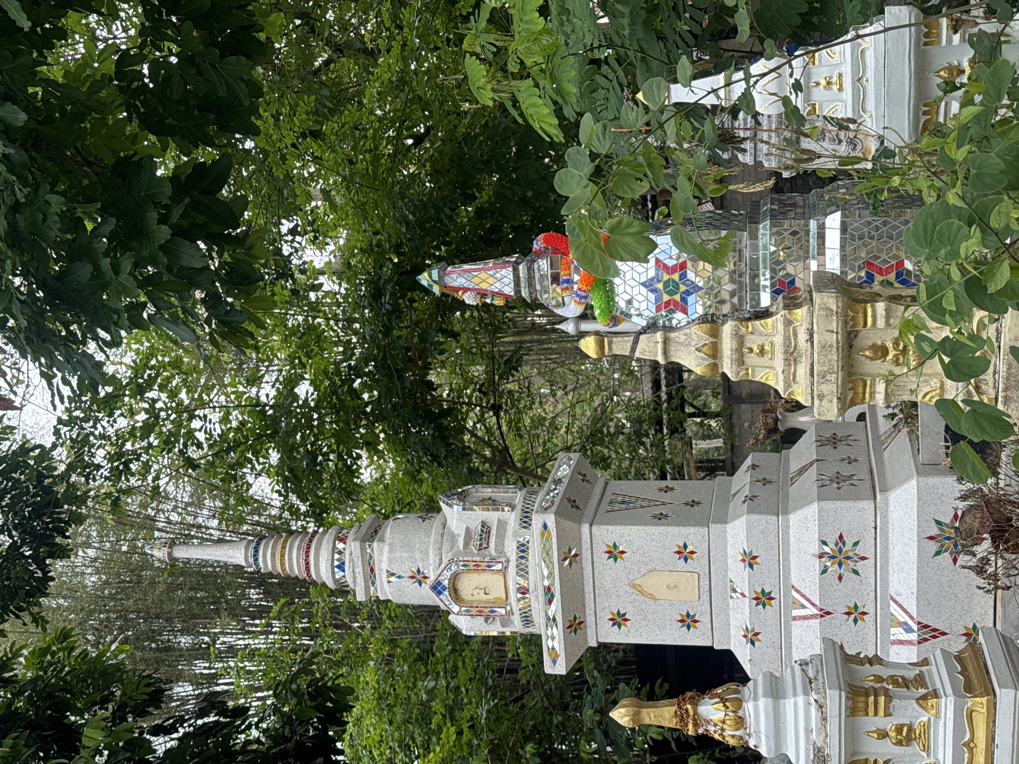 The image shows two religious monuments in an outdoor setting with lush greenery. One monument is white with colorful, star-shaped decorations. It has a tapering tower-like structure. The other monument is decorated with mirror mosaic and is adorned with colored garlands. The monuments are situated amongst trees and foliage. The location is Ban Sai Kham, Thailand.