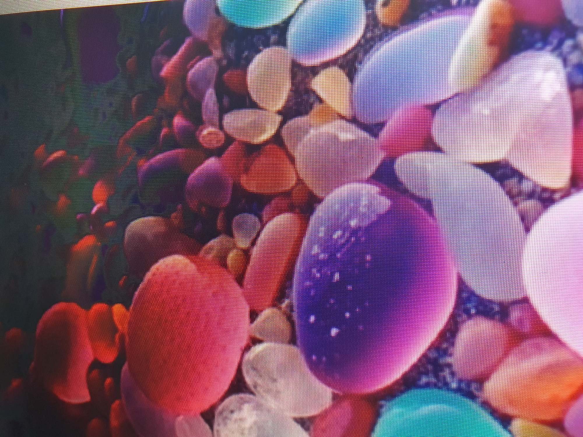 The image displays a close-up view of smooth, colorful pebbles. The pebbles are rounded and vary in size, color, and translucency. Colors include shades of red, pink, purple, blue, teal, orange, and white. Some pebbles appear to be translucent and have a glassy texture, while others are more opaque. The pebbles are densely packed together on a dark, granular surface, possibly sand or gravel. The lighting suggests the objects are illuminated from above, casting soft highlights on their surfaces and creating a sense of depth. The image's perspective is from directly above, looking down at the collection of pebbles. There are no visible people, animals, or any discernible elements that would link this scene to a specific location in Tashkent, Uzbekistan, or any other city. The image is purely focused on the abstract arrangement and coloration of the pebbles.