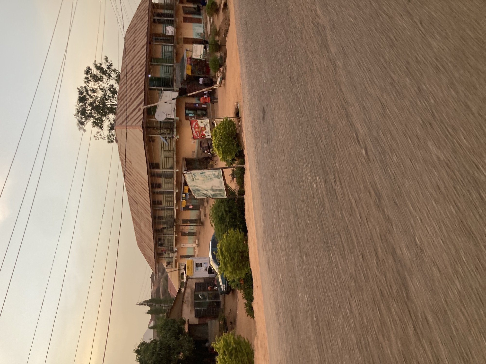 The image shows a street scene in Jos, Nigeria, viewed from the perspective of a moving vehicle. The road is the dominant element in the foreground, with a blurred texture suggesting motion. To the left of the road, a multi-story building with a corrugated metal roof stands prominently. The building has multiple windows and appears to house several businesses or residences, indicated by the various signs and objects visible in front of the individual units. Power lines are visible against the sky. Vegetation, including small trees and shrubs, grows along the edge of the road and in front of the building. A few people are present but indistinct.