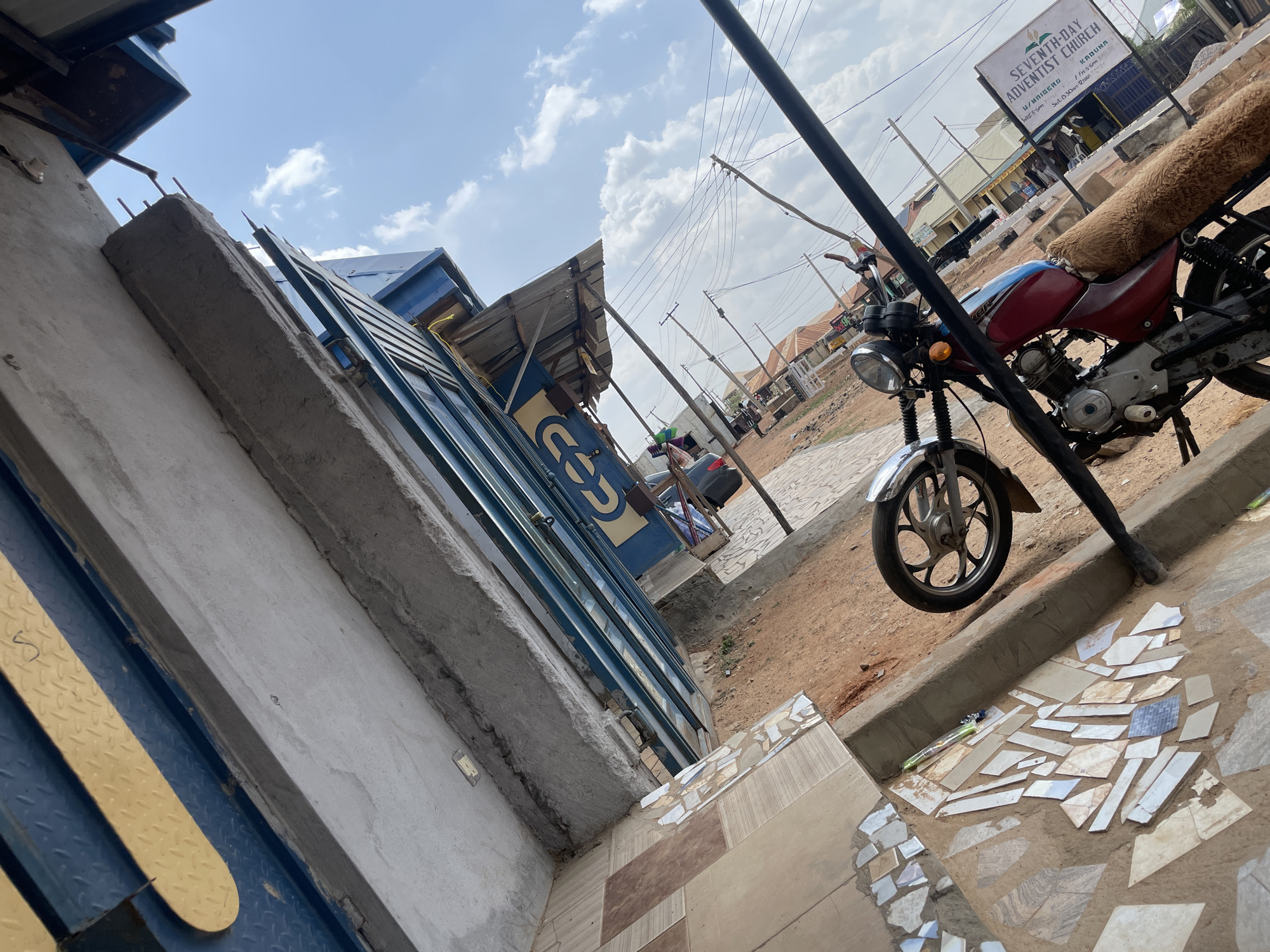 This image captures a bright, daytime street scene in Narayi, Nigeria, with a slightly tilted, low-angle perspective. In the immediate foreground, a rough concrete structure with exposed rebar extends upwards, alongside a yellow-patterned metal surface, possibly part of a gate or door. Adjacent to this, a blue building with a corrugated metal roof displays a partial sign with the letters "COD".
Across a stretch of ground comprising dirt, broken tiles, and patterned flooring, a red and blue motorcycle is parked, partially obscured by a black pole. The motorcycle features a distinctive brown, shaggy seat cover. Its front wheel and headlight are clearly visible.
In the background, a prominent white sign for the "SEVENTH-DAY ADVENTIST CHURCH" is visible, detailing its location as "U/MAIGERO, KADUNA" and listing service times: "Wed: 5-6pm", "Fri: 5-6pm", and "Sat: 8:30-12noon". The street beyond the motorcycle is characterized by multiple utility poles and overhead power lines, with other buildings lining the thoroughfare. The sky is clear blue with scattered white clouds, indicating good weather. No specific activity or people are depicted; the scene is a static snapshot of a typical roadside environment.