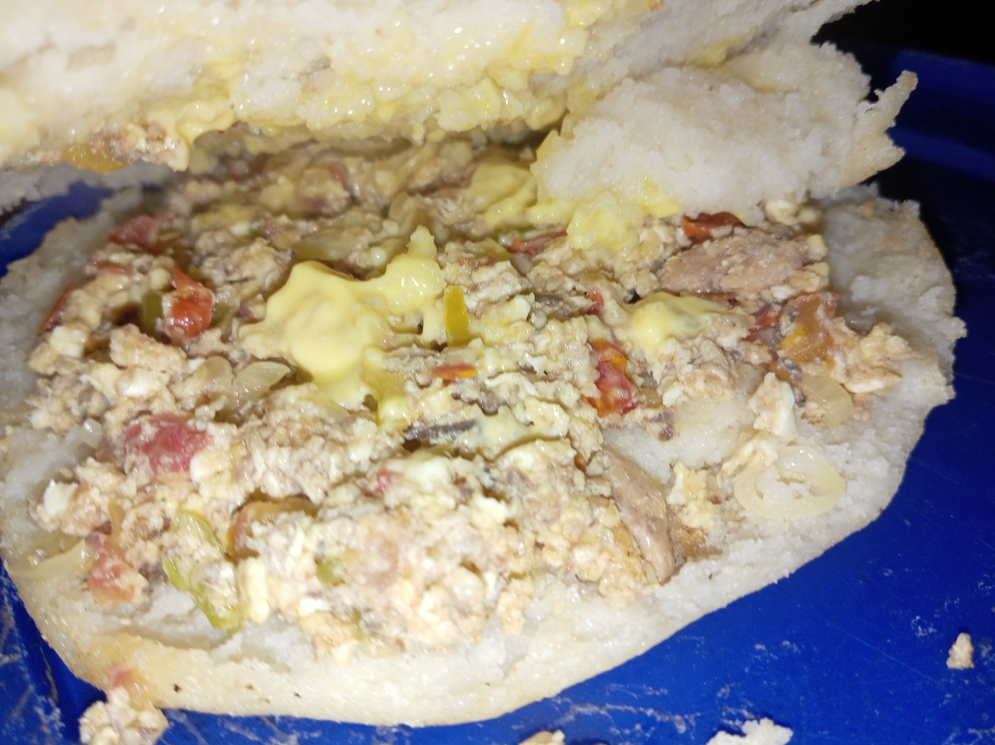 A close-up shot depicts an arepa filled with scrambled eggs, possibly tuna, tomatoes, peppers, onions, and yellow sauce served on a blue plate. The dish is in Parroquia Ocumare, Venezuela.