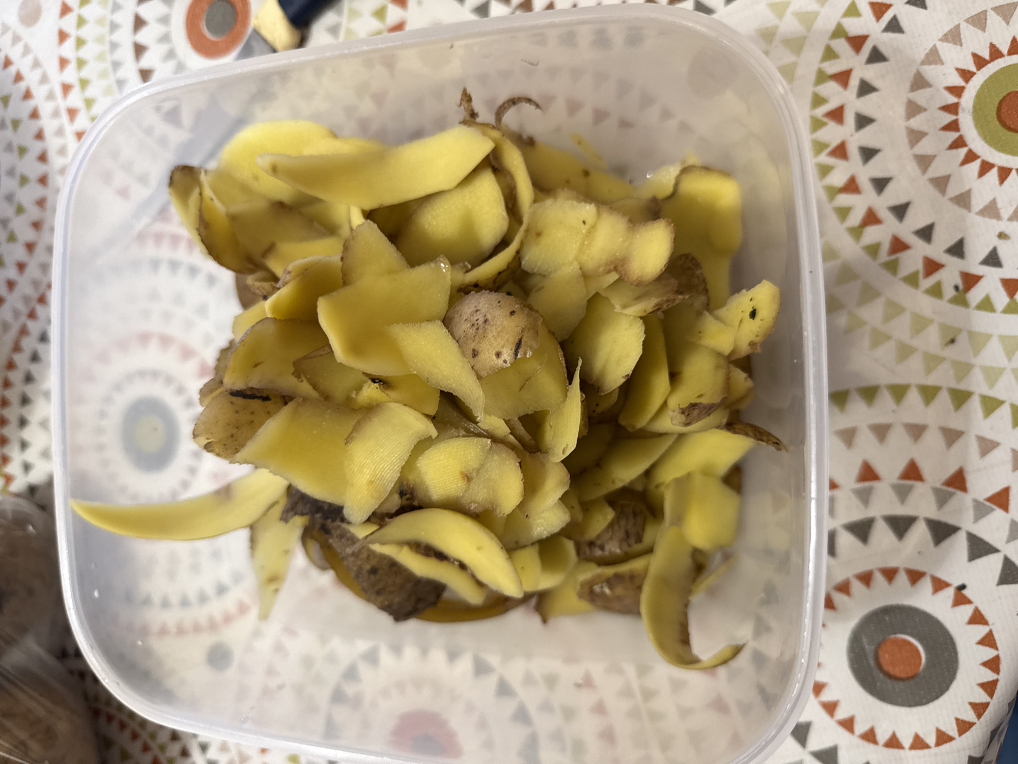 A clear plastic container filled with potato peels is placed on a table. The container is positioned centrally in the frame, and the peels are piled high within it. The potato peels are mostly yellow with some brown skin still attached, indicating they have been recently peeled. Some of the peels are curled, while others are laid flat. The table beneath the container has a patterned tablecloth featuring circles and triangles in various colors, including red, orange, grey, and green. The background is slightly out of focus, but a blurry object wrapped in plastic, possibly more potatoes, can be seen in the upper left corner. A portion of a knife with a blue handle is visible on the right side of the frame, resting on the tablecloth. The lighting appears to be natural, casting subtle shadows on the peels. The image does not provide any specific indicators of a location in the city of Морозовка, Russia.