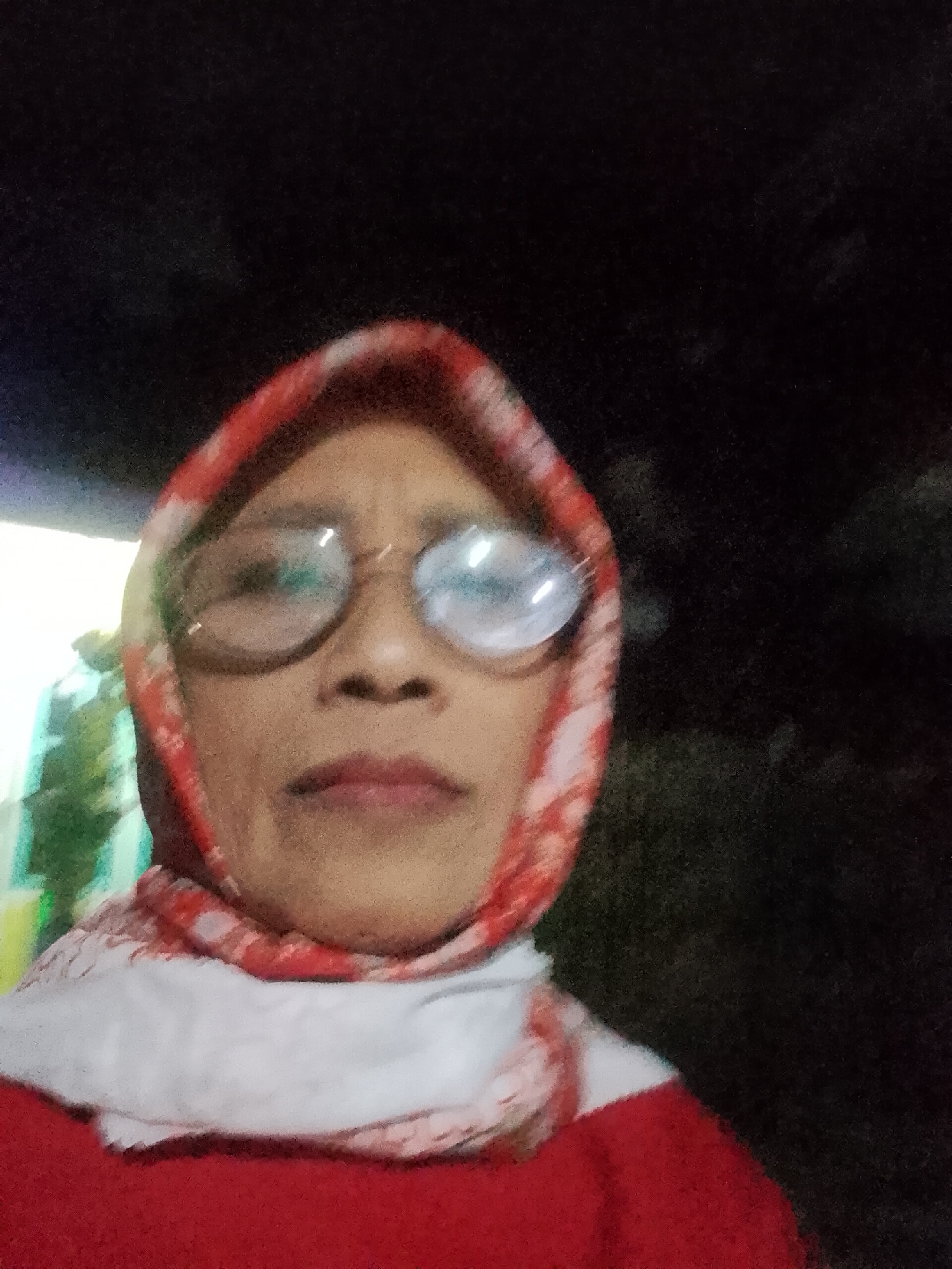 A selfie of a woman wearing a red hijab and glasses is captured in a dimly lit environment. She is wearing a red top and a white scarf. The background is dark and out of focus, suggesting it is nighttime. The woman's face is centered in the frame, and her expression is neutral. No specific location within Kecamatan Pati, Indonesia can be identified from the image.