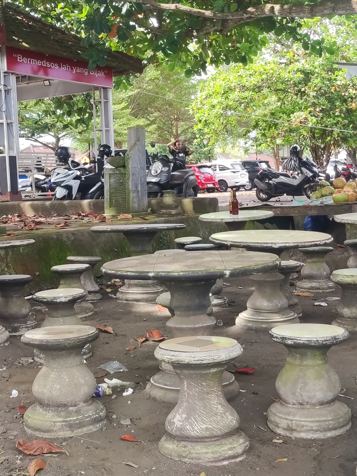 The image depicts an outdoor area with numerous round concrete tables and stools, some with tiled tops, set on a dirt ground covered with scattered leaves and litter. In the background, there are several motorcycles parked near a raised concrete platform that supports a structure with a sign that reads "Bermedsos lah yang Bijak". Behind the motorcycles are parked cars. A person wearing a helmet is sitting on a motorcycle. On one of the tables is a brown bottle and a pile of coconuts. A man is adjusting his hair near the parked motorcycles. The environment is shaded by trees. The location is in Padangbai, Indonesia.