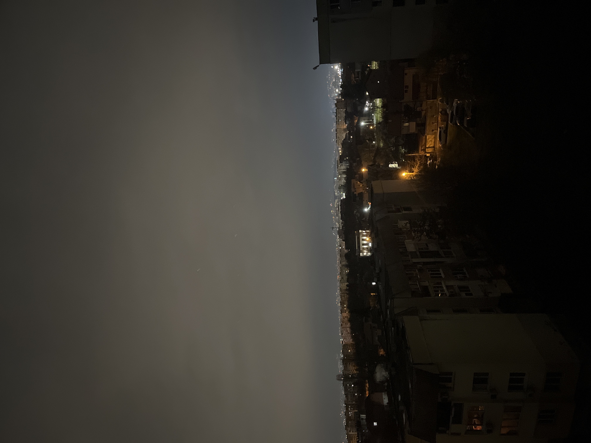 The image shows a nighttime view of Sofia, Bulgaria. The left side of the image depicts a hazy sky. The center shows the city skyline with various buildings and streetlights. Buildings are visible on the right side of the image. The environment is dark, suggesting the photo was taken at night.