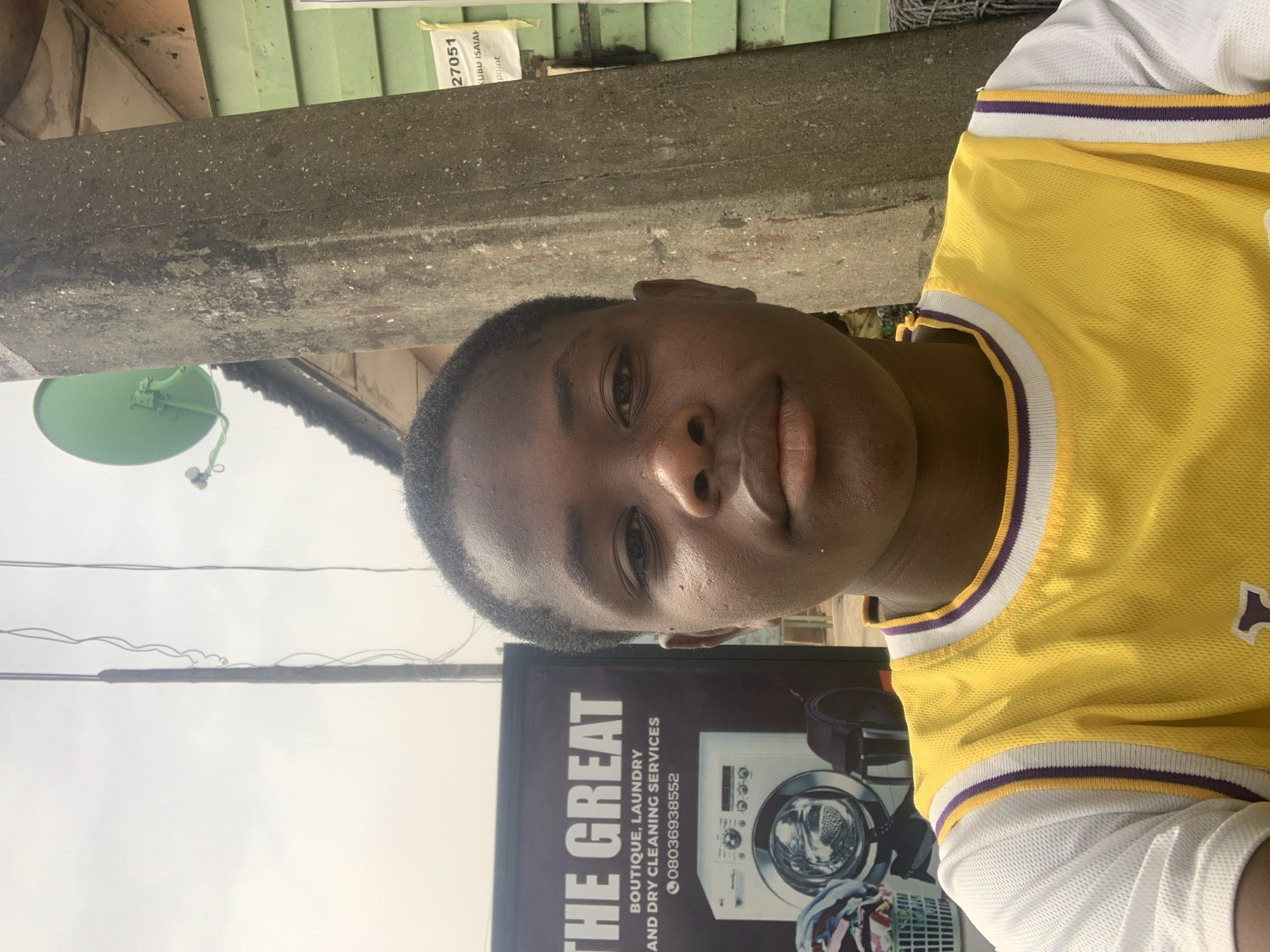 A young person with dark skin and short dark hair looks directly at the camera with a neutral expression. They are wearing a yellow sleeveless sports jersey with purple and white trim, possibly a basketball jersey, over a white short-sleeved undershirt.
The setting appears to be an urban outdoor environment. Behind the person, a rough, grey concrete beam or ledge runs horizontally. Above this beam, a light green corrugated metal structure is visible, along with a small, partially visible white paper or sign bearing the text "27051", "KUBU ISAIAH", and "Point". To the left of the person's head, a green, circular, dish-like object with an attached antenna is visible.
To the right and slightly behind the person, a large dark sign advertises "THE GREAT BOUTIQUE, LAUNDRY AND DRY CLEANING SERVICES". The sign also displays the phone number "08036938552" and features an image of a washing machine. Overhead, several electrical wires are strung across the sky. The bright lighting suggests it is daytime. The overall scene indicates a commercial or residential street in an urban area, consistent with Jos, Nigeria.