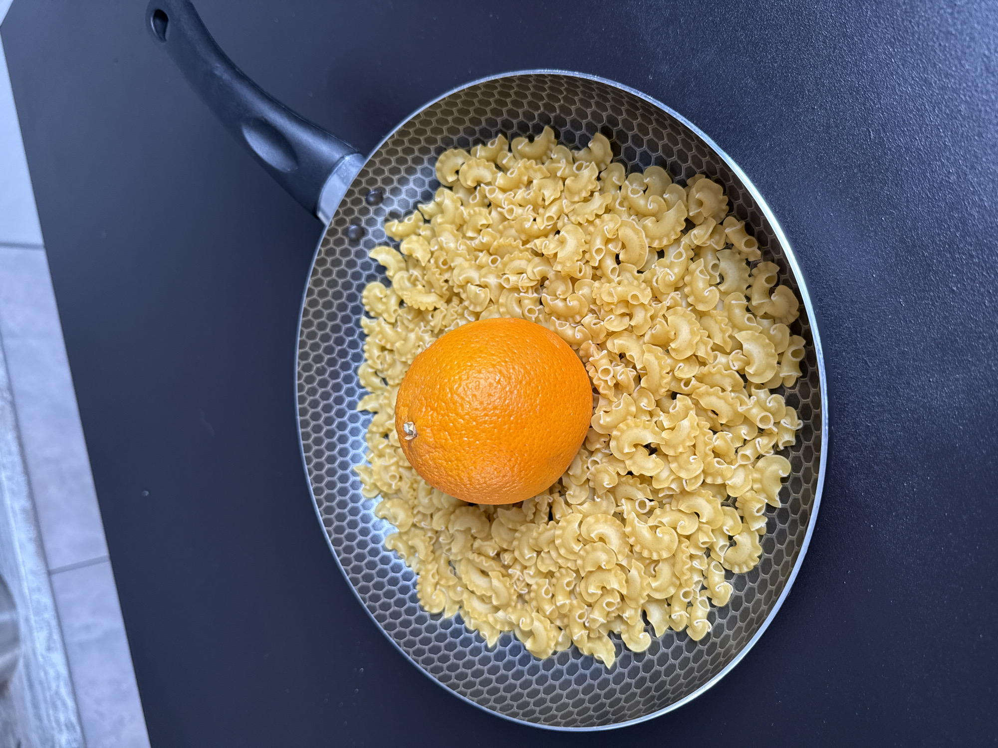 A top-down shot shows a frying pan filled with elbow macaroni and an orange in the center. The pan has a black handle and a light gray interior with a hexagonal pattern. The macaroni is pale yellow. The orange is bright orange and intact. The pan sits on a dark gray surface, with a sliver of light gray surface visible in the upper left corner. The image does not contain information to determine the location in the city of Rostov-on-Don, Russia.