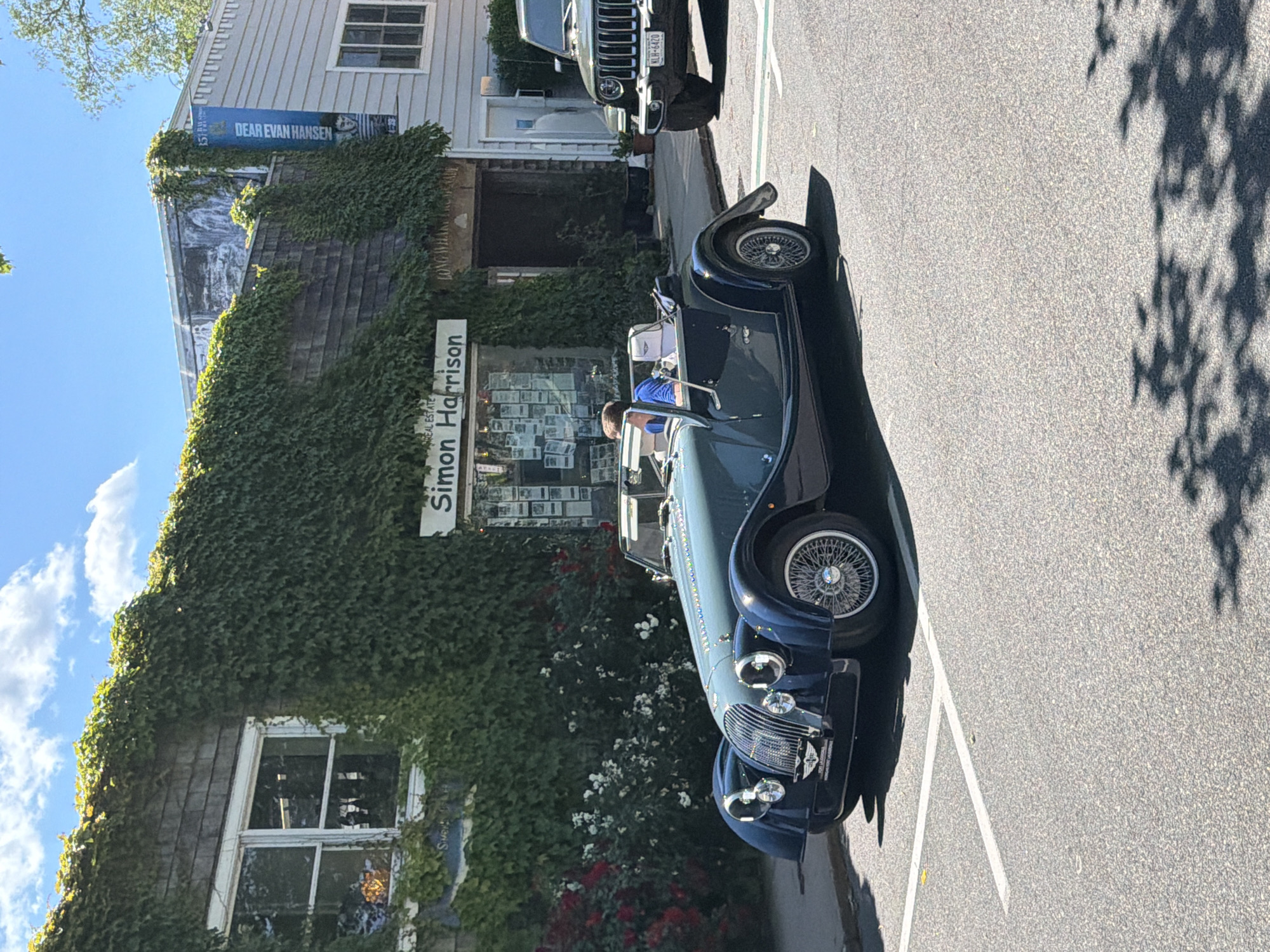 A dark blue convertible classic car is parked on a street in the Village of Sag Harbor, USA. The car has wire-spoke wheels and a prominent grille. A person wearing a blue shirt is seated in the driver's seat. Behind the car, a building is covered in dense green ivy, with a sign that reads "Simon Harrison" above a shop window. To the right of the building, another sign hangs vertically and reads "DEAR EVAN HANSEN". Further to the right, the rear of a dark green Jeep Wrangler is visible, with a license plate that reads "KLH-6420". The scene is set on a sunny day with a bright blue sky and scattered clouds.