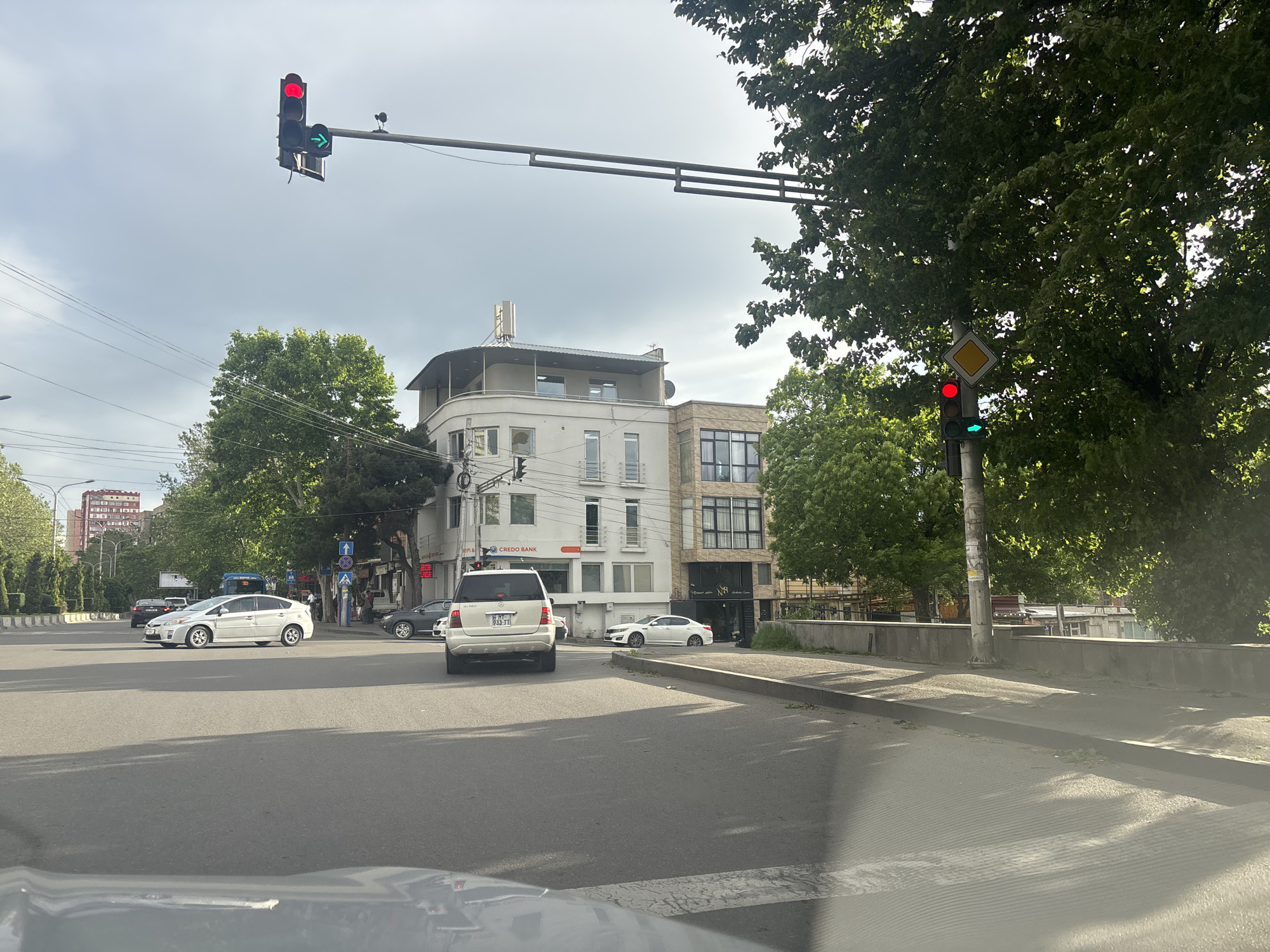 The image depicts a street scene in Tbilisi, Georgia. The view is from inside a car, looking out onto an intersection. A white Toyota Prius is visible in the left lane, and a white Mercedes SUV is in the center lane. Both vehicles are approaching the intersection, where the traffic signals indicate a green arrow for turning and a red light for going straight. A Credo Bank building is on the corner. The overall environment suggests an urban area on a partially sunny day.