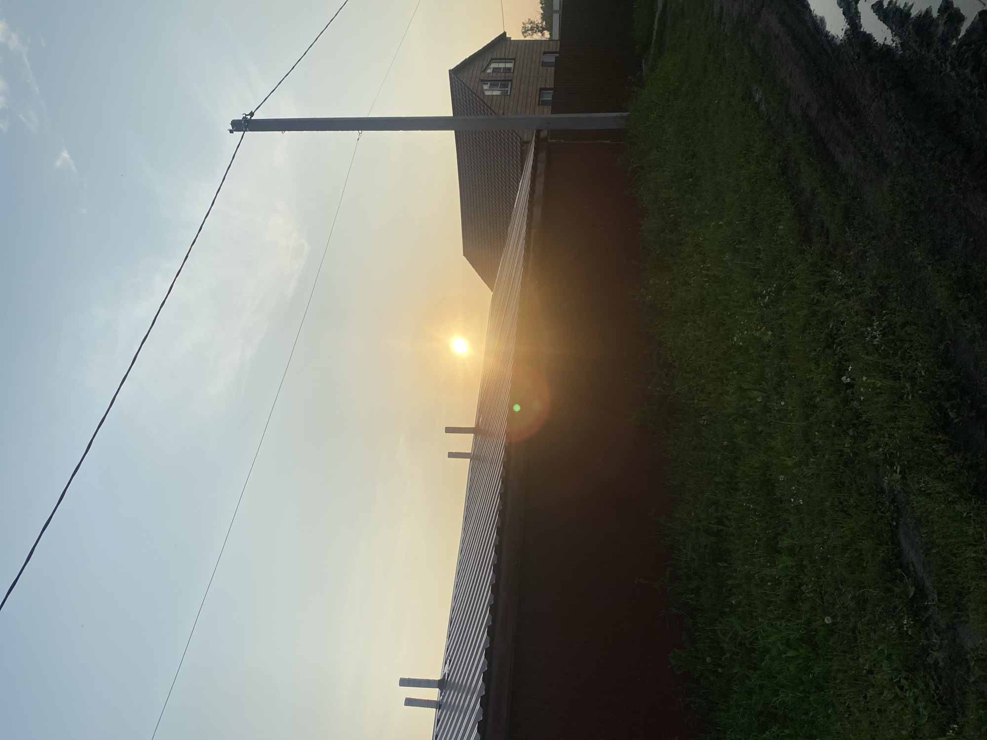 The image captures a suburban or rural scene in Verkhny Karbush, Russia, during what appears to be either sunrise or sunset. The sun, a bright orb, is positioned in the upper left quadrant, casting a warm glow across the sky. In the foreground, a long, dark red building with a corrugated metal roof dominates the left side of the frame. Several ventilation pipes protrude from the roof. To the right of this building, a two-story wooden house with a dark-tiled roof is visible, featuring multiple windows. A dark fence runs behind the wooden house. A utility pole with wires stretches diagonally across the sky, and another wire runs parallel to it. The ground in front of the buildings is covered in lush green grass, with some patches of mud and puddles of water on the right side of the frame, indicating recent rain or a damp area. The overall atmosphere is tranquil, with the soft light creating a serene ambiance. No people are present in the image.