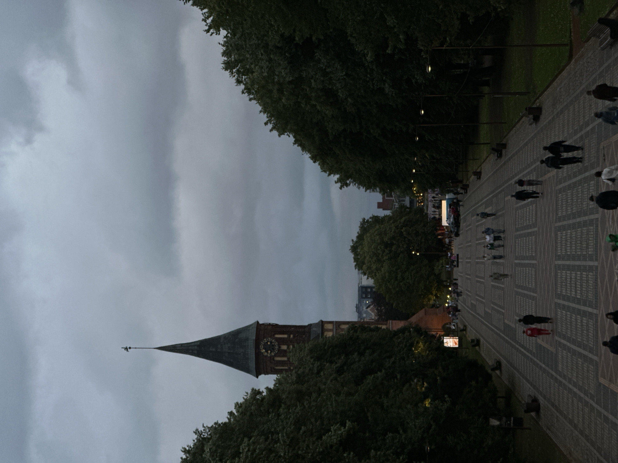 The image depicts a wide, paved pedestrian walkway lined with trees on both sides, leading towards a large, brick clock tower with a pointed spire. The sky is overcast and dark, suggesting dusk or an impending storm. Several people are scattered along the walkway, some walking in groups, others individually. In the foreground, a few people are visible, with the most prominent being a couple in red and black attire walking away from the camera. Further up the path, groups of people can be seen strolling, and a vehicle is parked on the side. The clock tower, identifiable as the Kaliningrad Cathedral (also known as Königsberg Cathedral), stands prominently in the background, its clock face visible. The overall atmosphere is somber due to the low light and cloudy sky. The architectural style of the tower and the presence of the cathedral clearly indicate the location as Kaliningrad, Russia.