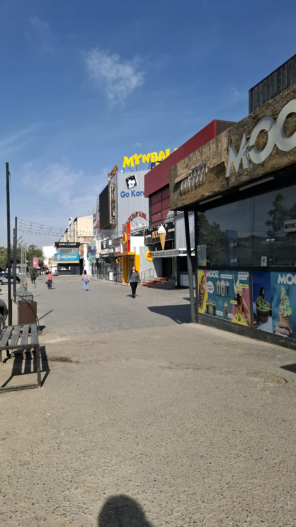 The image depicts a street scene in Shymkent, Kazakhstan, likely a commercial area. The foreground shows a paved pedestrian walkway with some debris and a shadow of a person. To the left, a metal bench is visible. The background features a row of shops and businesses with various signage. Notable signs include "MYNBALA", "Go Korea", "WE DRINK", and "MOOZ" with images of ice cream. Several people are walking along the street, some individually and others in small groups. The sky is blue with scattered clouds. The architecture appears to be modern, with a mix of storefronts, some with large glass windows displaying products. The overall impression is a public space on a clear day, likely during daytime hours.