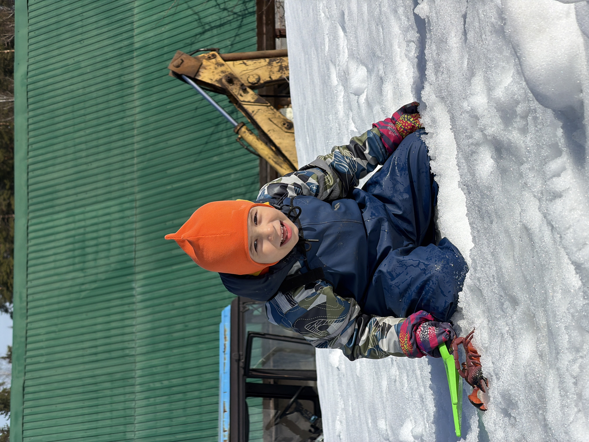 A young child wearing an orange hat, a blue snowsuit, and patterned gloves sits in the snow. The child is holding a toy crab and a green plastic tool. Behind the child, a green corrugated metal building is visible. A yellow and black piece of heavy machinery, possibly a backhoe, is positioned near the building. The location is possibly Chaikovsky, Russia, based on given instructions.