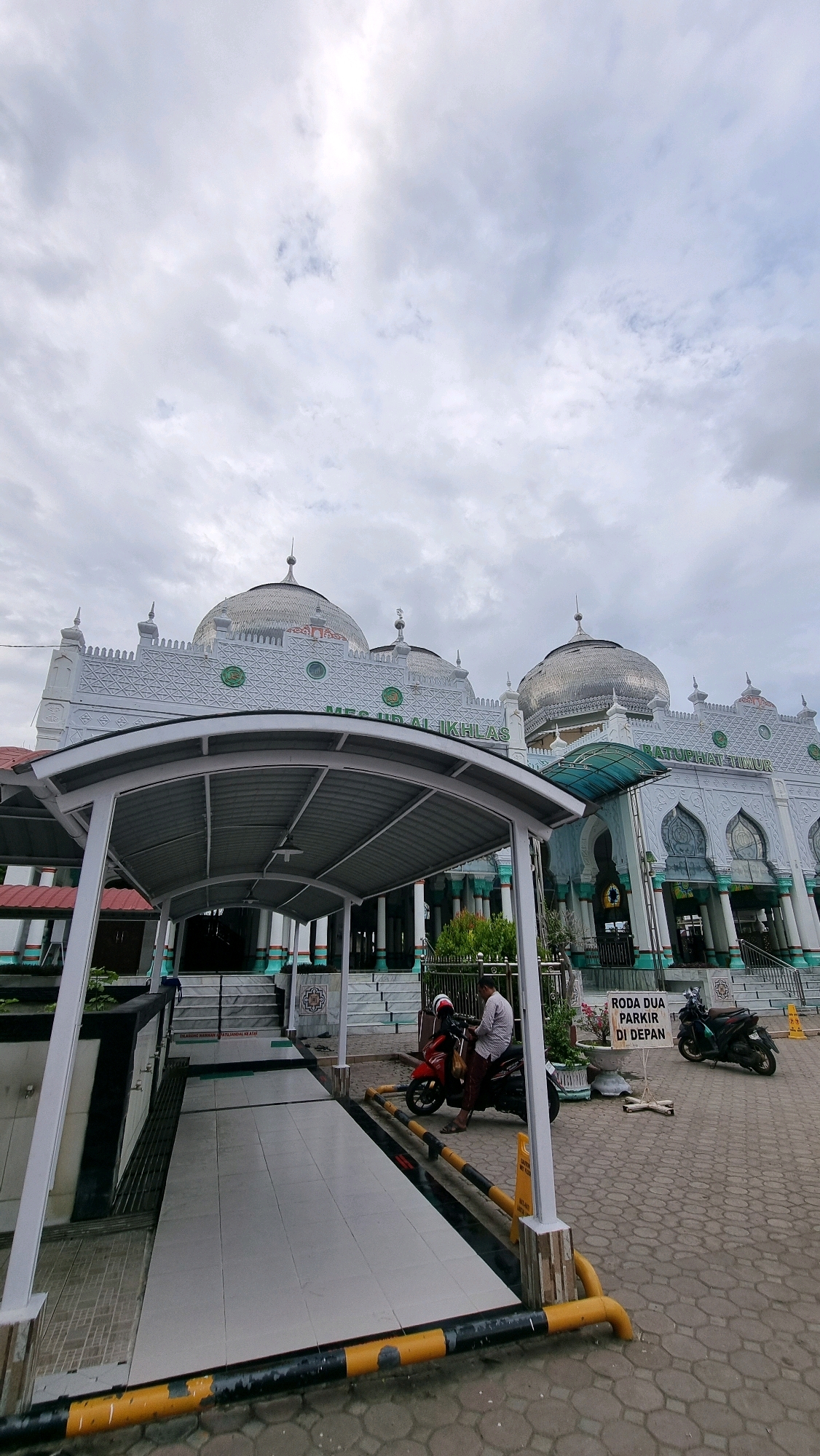 The image shows the exterior of a mosque in Lhokseumawe, Indonesia, under a cloudy sky. The mosque features white walls with intricate patterns, multiple domes, and green accents. In the foreground, there is a covered walkway with a tiled floor and yellow and black striped barriers. A red motorcycle is parked to the right of the walkway, with a person wearing a light-colored shirt and dark pants sitting on it. Another motorcycle is parked further to the right, and a sign with Indonesian text is visible behind the red motorcycle. A few potted plants are also present in the scene. The ground is paved with interlocking stones.