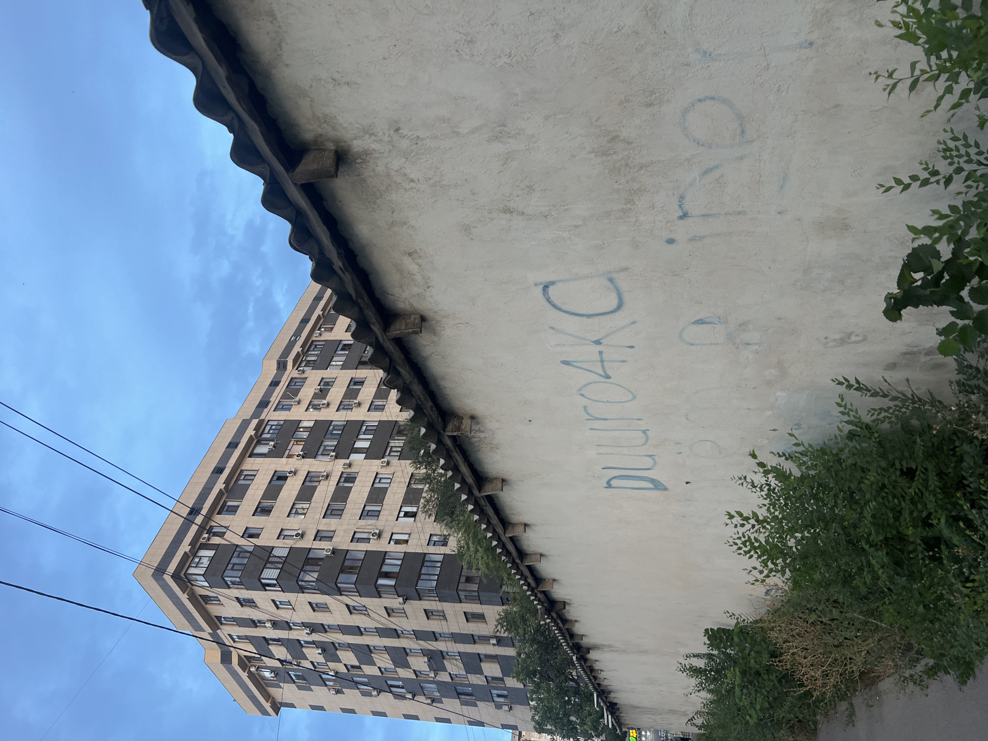 A light beige, textured concrete wall with a slightly wavy, dark tiled roof runs diagonally from the bottom right to the top left of the frame. Blue graffiti is visible on the wall. In the upper left quadrant, a tall, modern apartment building with a checkered facade of beige and dark grey panels and many windows is visible against a blue sky with scattered clouds. Green foliage lines the bottom of the wall and the lower left corner of the image. Power lines crisscross the sky above the apartment building. The scene appears to be in an urban environment.