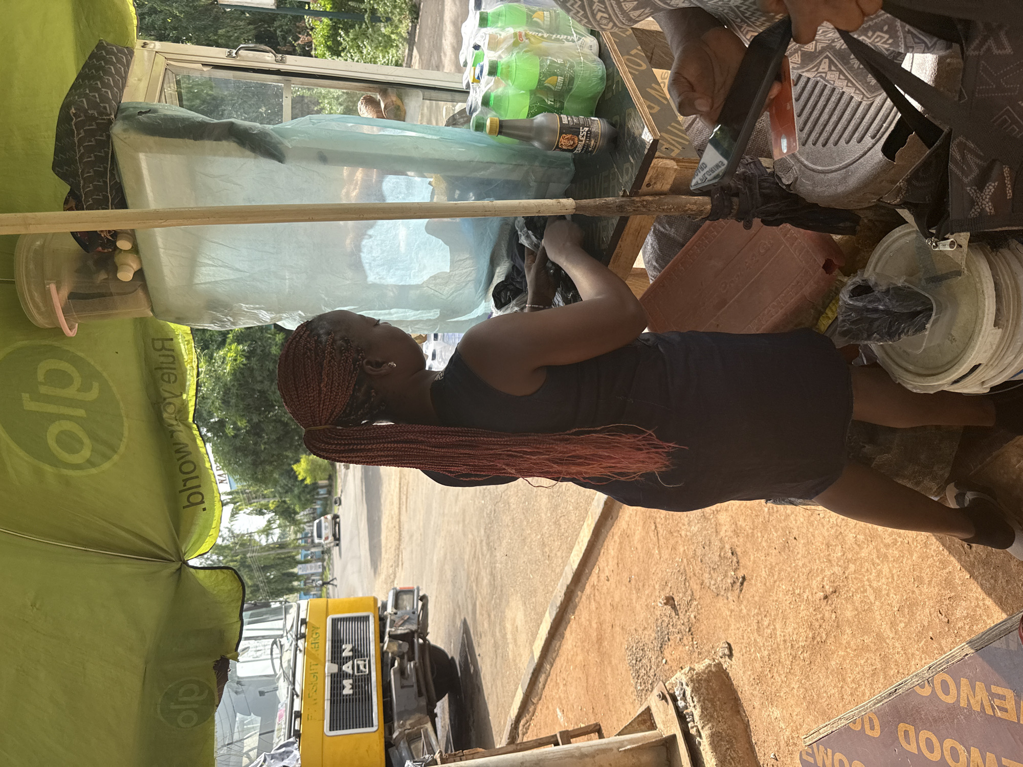 This image captures an outdoor street vendor scene, likely a roadside stall, in Abuja, Nigeria. The main subject is a woman with long, reddish-brown box braids, viewed from her right side, facing towards the left of the frame. She wears a dark top and appears to be attending to the stall, with her hands engaged in an activity on the surface before her. Another person's hands are partially visible in the upper right quadrant, possibly interacting with items on the stall or holding a mobile phone.
The stall consists of a makeshift counter, possibly made of wood. Various items are displayed for sale or use. To the left, a large light blue plastic covering likely conceals a cooler or storage unit, and a clear plastic container with a pink handle sits nearby. In the center, a pack of green bottled soft drinks is visible, alongside a single dark brown bottle labeled "FEARLESS." The stall is partially shaded by a large green umbrella or canopy with text "alo" and ".bhow.move" (reversed) printed on it.
The ground is a mix of reddish-brown dirt and some paved areas. In the background, a yellow truck with "MAN" visible on its grille and "FARSIGHT ENERGY" (reversed) on its side is parked on the street. Trees and foliage line the background, suggesting a sunny daytime setting. Other vehicles are faintly visible in the distance. Additional objects include a red plastic crate or tray with "Cool Kid" (reversed) partly visible, stacked white plastic lids or bowls, and a wooden board in the lower right corner displaying "WOOD O" and "HOMEWO" (reversed). The scene depicts everyday commerce in a bustling outdoor environment.
