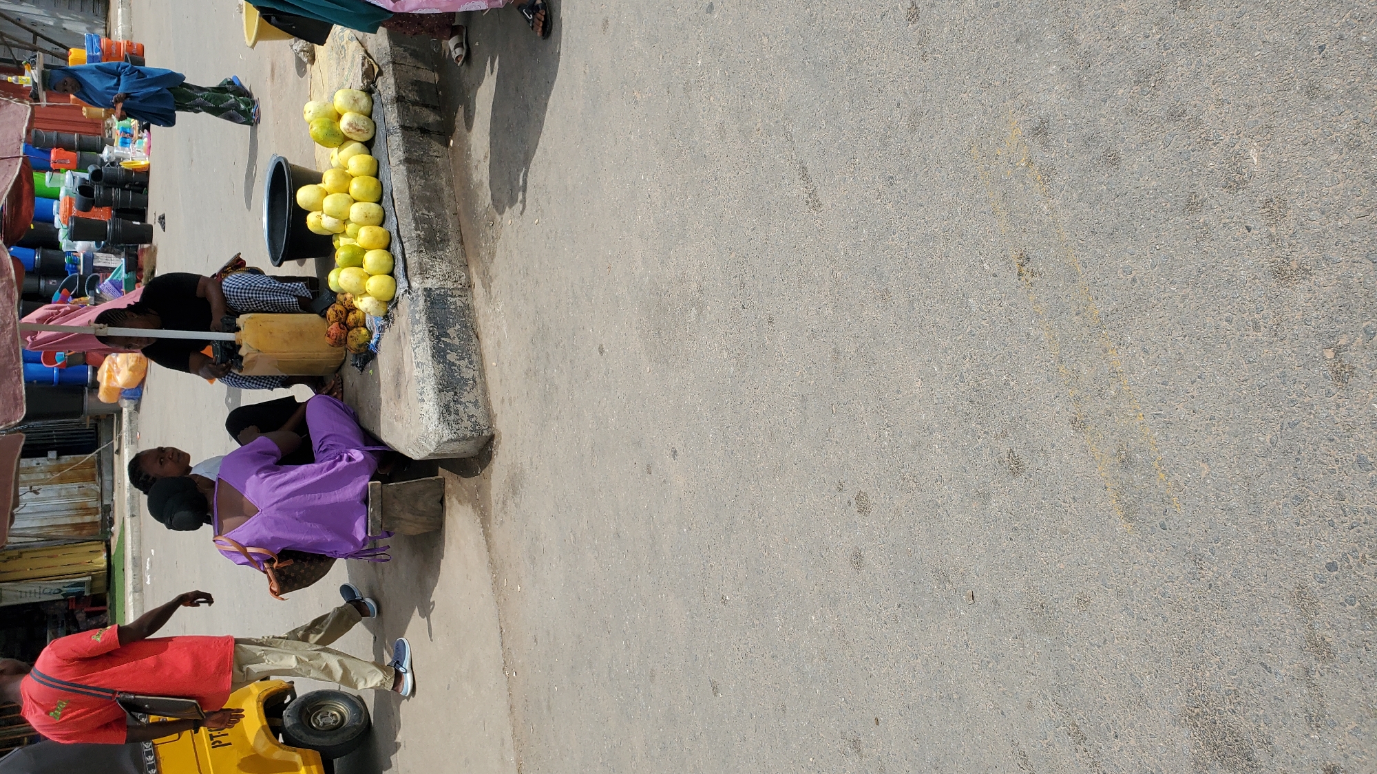 In Jalingo, Nigeria, on a sunny day, a street scene unfolds near a market. A man in a red t-shirt walks away from a yellow three-wheeled vehicle. A woman in a purple dress sits on a low wooden stool, facing away from the camera. Another woman is seated beside her, partially visible. In the background, a woman in a black shirt and checkered skirt sits behind a yellow jug, with a pile of fruits, possibly mangoes, displayed in front of her. Several large plastic buckets and other market goods are visible behind the stall. Further in the background, a woman in a blue headscarf and patterned skirt walks away from the market. The ground is asphalt, with a curb visible in the foreground.